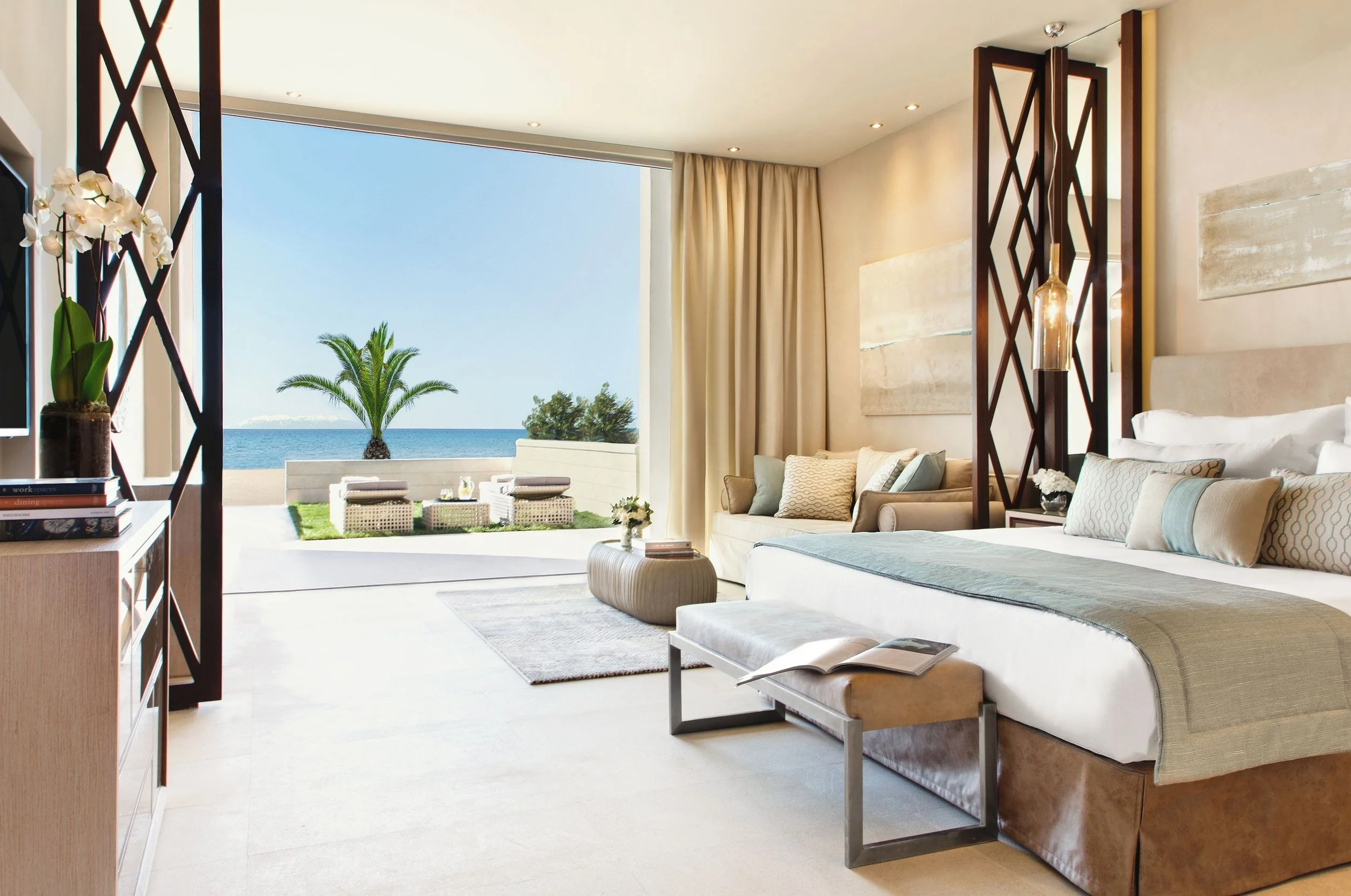 A living room with a view of a beach and ocean, featuring beige sofas, a small round beige ottoman, a bookshelf with books and a potted orchid, and large windows with beige curtains.
