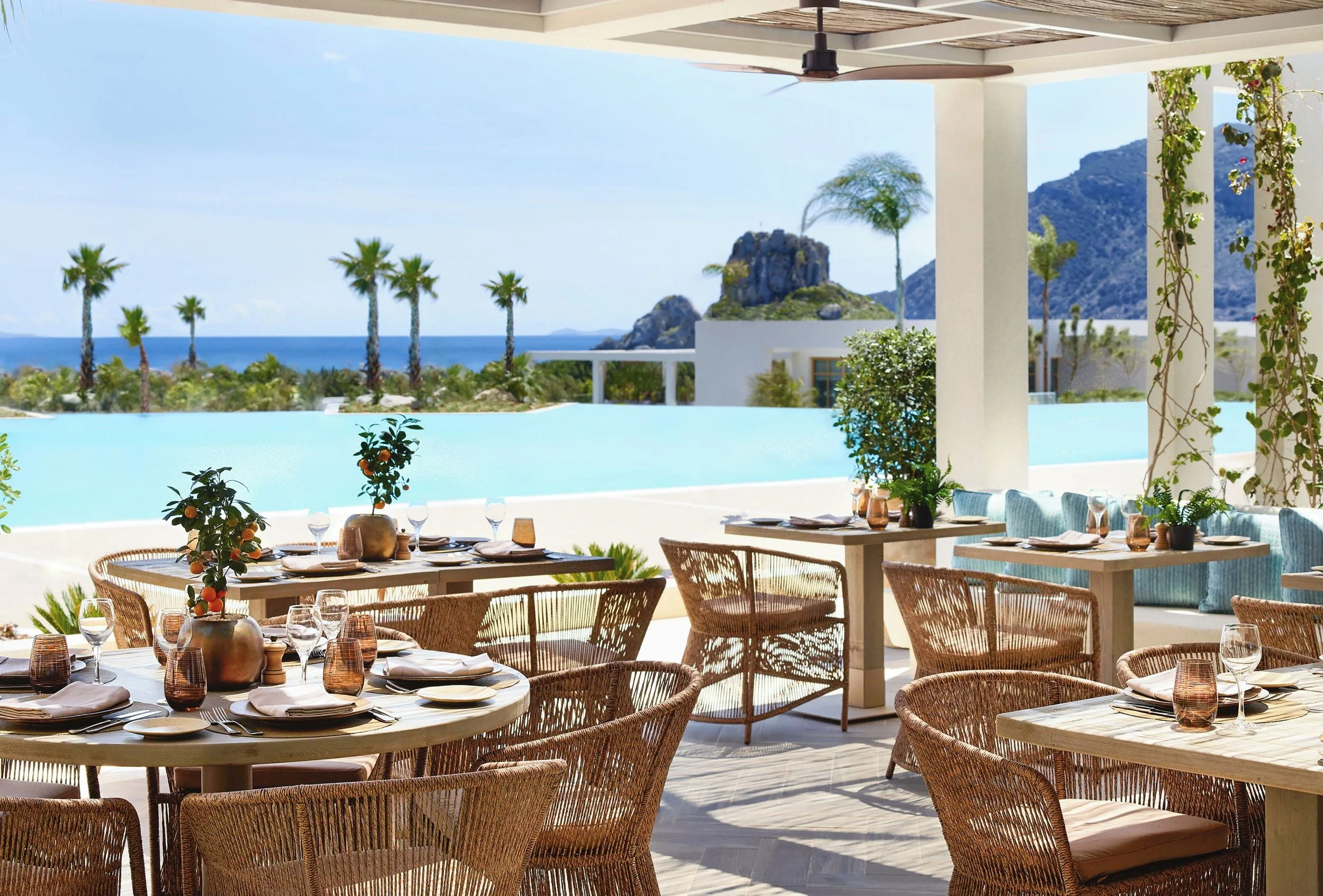 Outdoor restaurant with set tables and chairs overlooking a scenic view of palm trees, mountains, and the ocean under a covered patio.