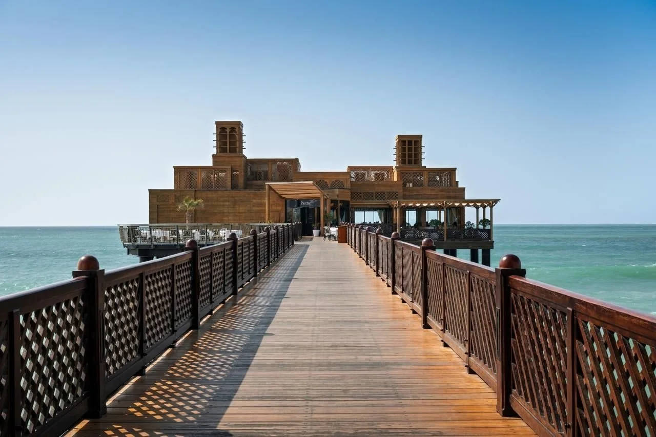 Wooden pier leading to a building with open-air balconies, situated on the ocean with blue sky overhead.