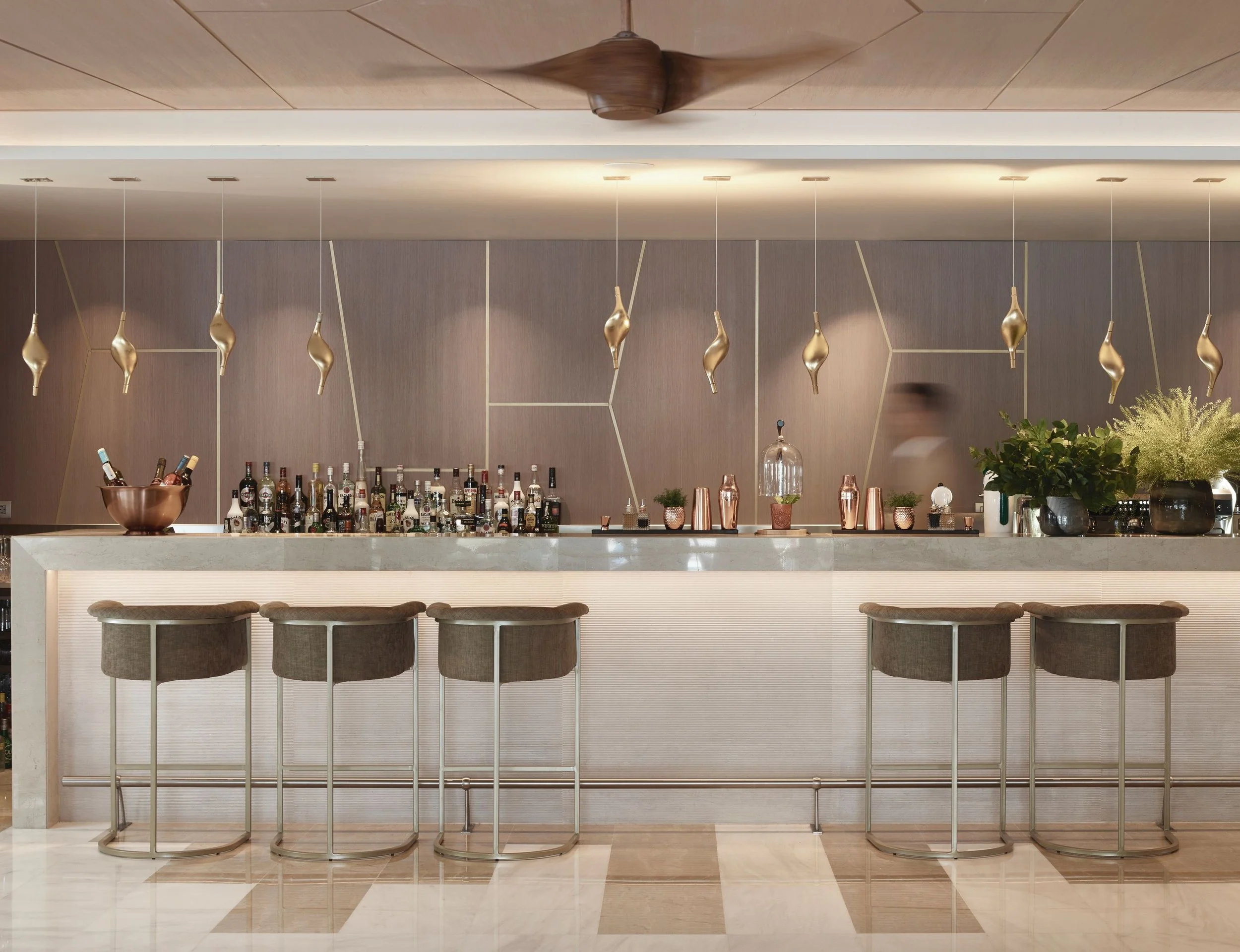 Interior of a modern bar with a marble counter, gold decorative pendant lights hanging from the ceiling, and a backdrop of shelves with various bottles and plants.