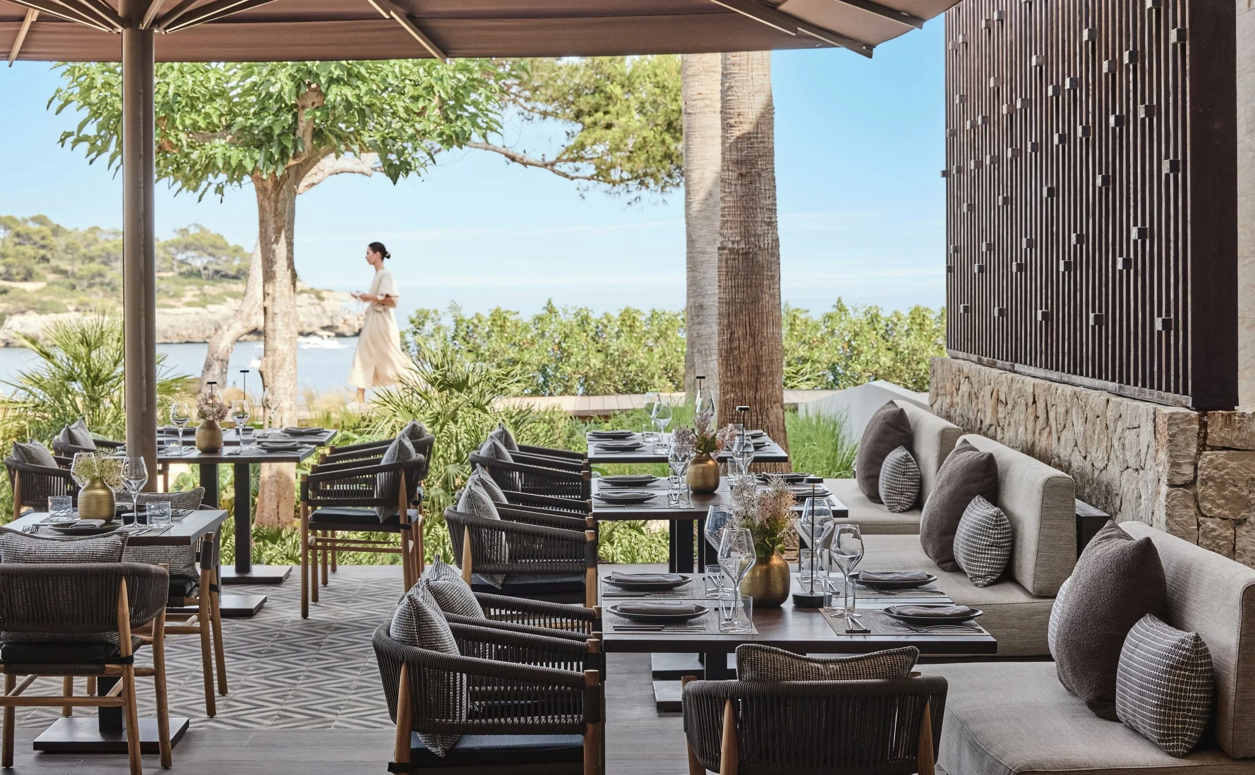 Elegant outdoor restaurant with tables set with wine glasses, plates, and cutlery, overlooking a scenic view with trees and water, shaded by umbrellas.