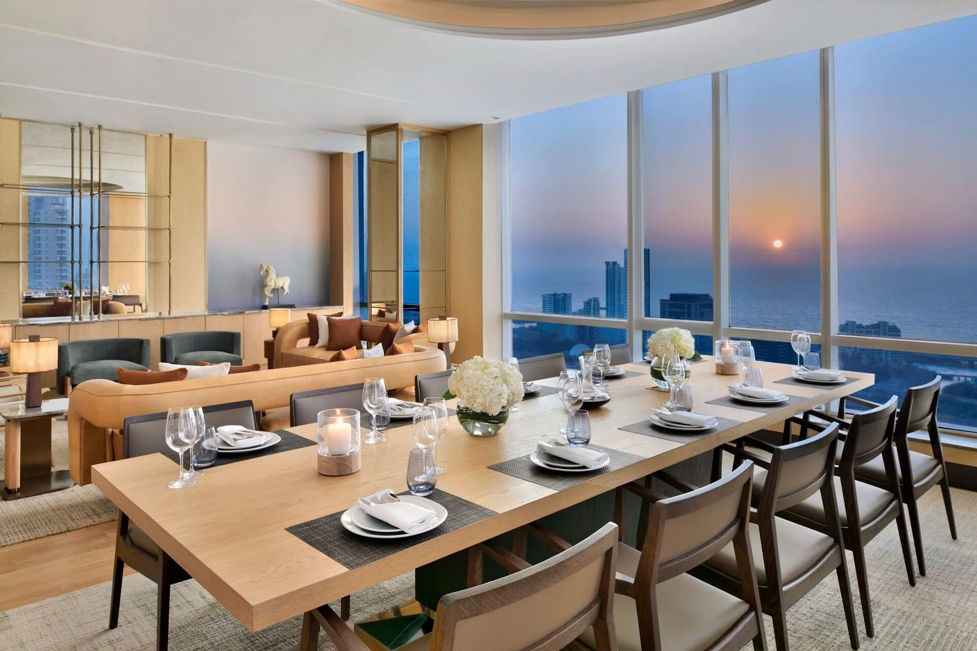 Modern dining room with large floor-to-ceiling windows showing a city skyline and ocean at sunset. The room has a long wooden table set for a meal with wine glasses, plates, and candles, and is decorated with white floral centerpieces.