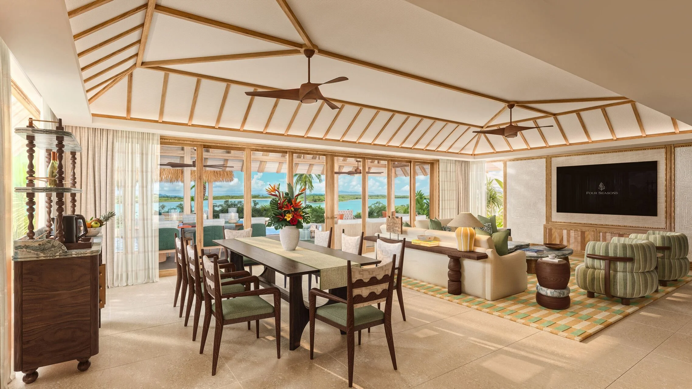 Sunlit living room with wooden ceiling beams and ceiling fans, overlooking a water view through large glass windows, decorated with tropical furniture and flowers.