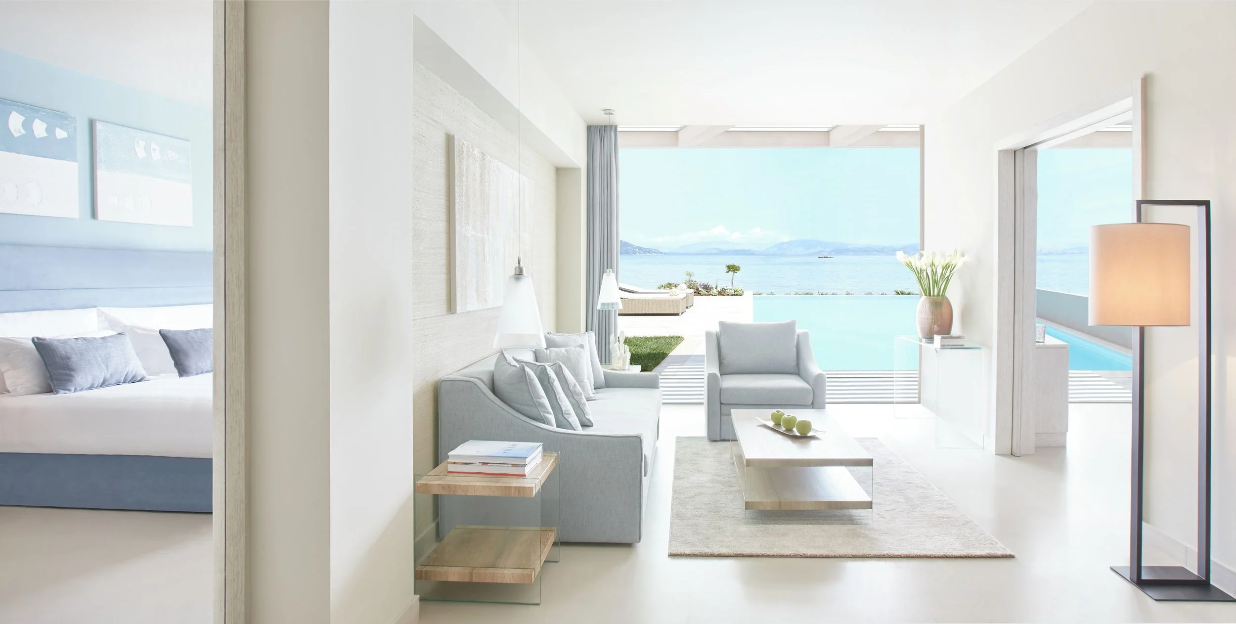 Bright modern living room with white and gray furniture overlooking a body of water and distant mountains through large glass windows.