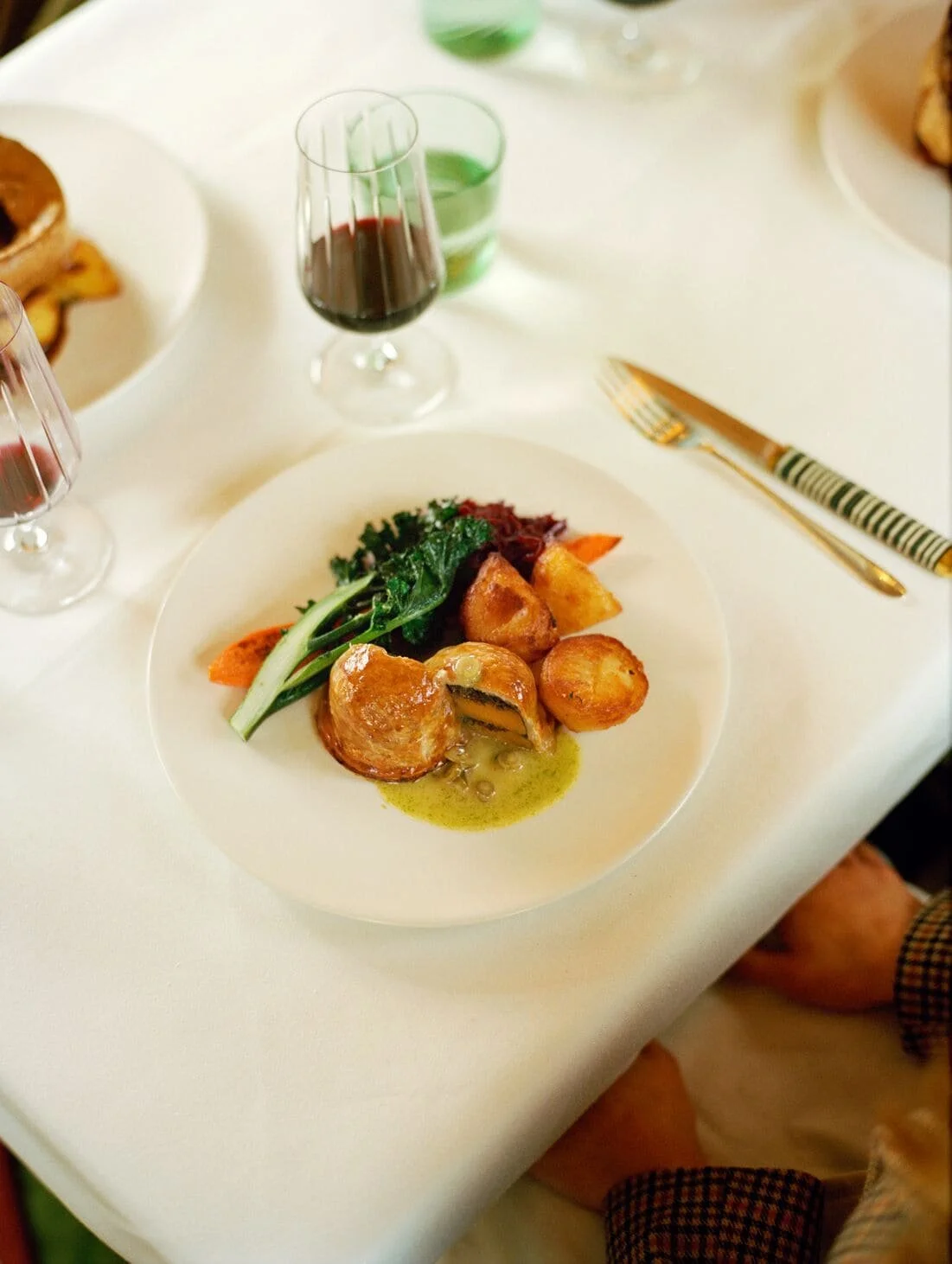 A fine dining plate with roasted vegetables, greens, and a sauce, with glasses of red and green drinks on a white tablecloth.