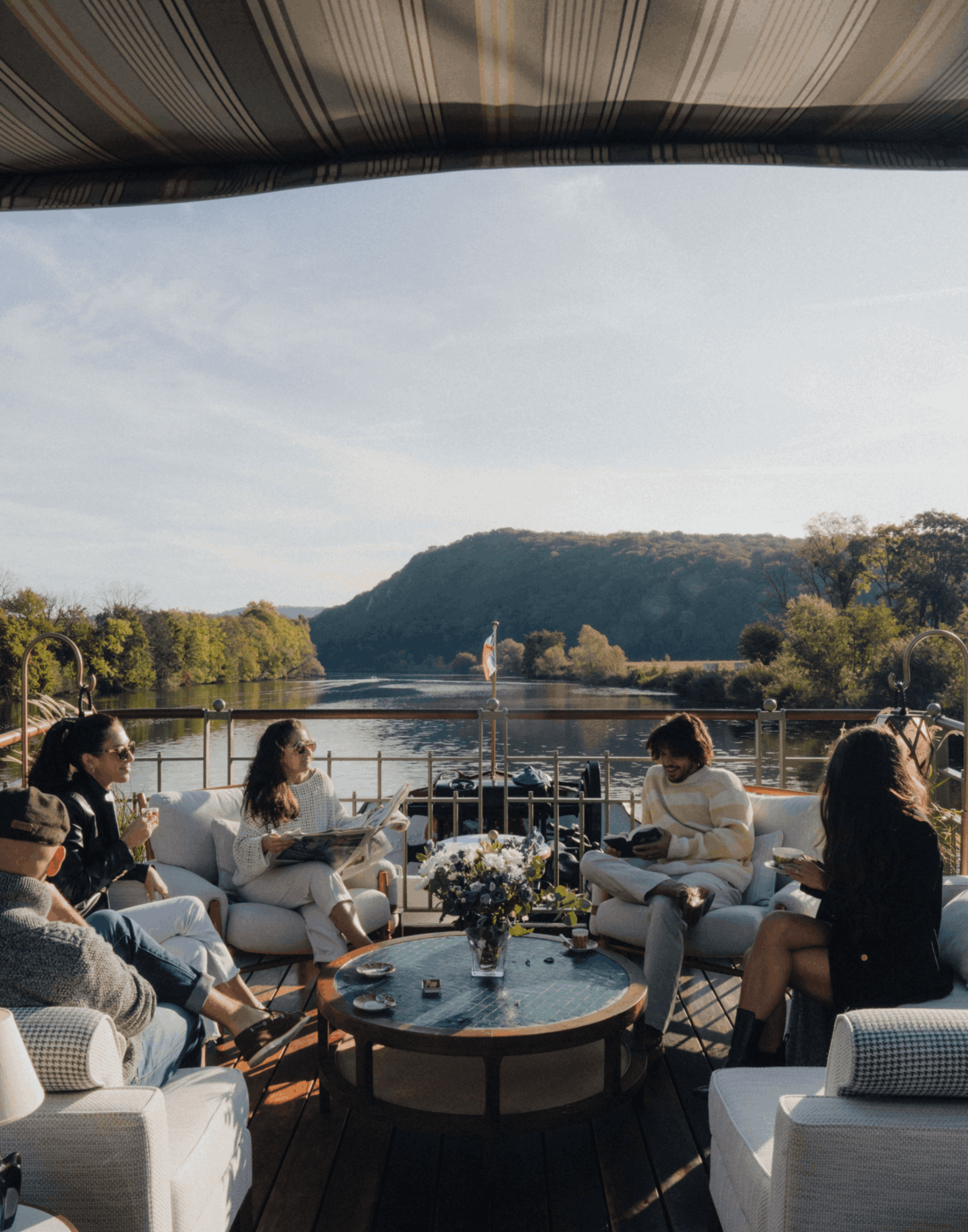 People sitting on outdoor patio furniture near a river, with trees and hills in the background, under a striped awning during late afternoon or early evening.