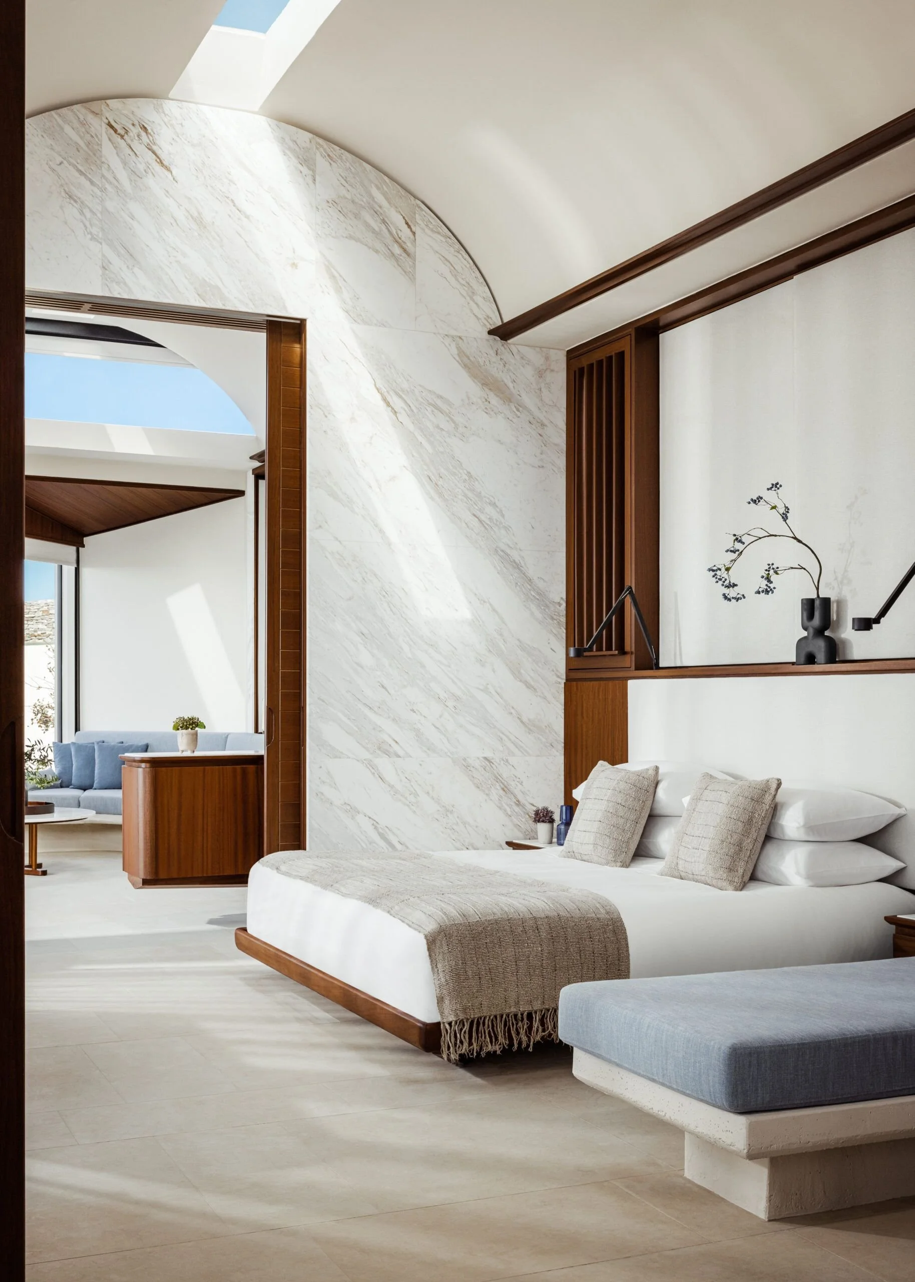 A modern, minimalistic living space with a white upholstered bed with beige throw blanket and square cushions, a gray upholstered bench, and a white textured floor. The room features a wooden accent wall with a built-in shelf holding a dark vase with a plant, and a section of large marble wall with beige veining. Two large windows allow natural light, with small potted plants visible on the side.