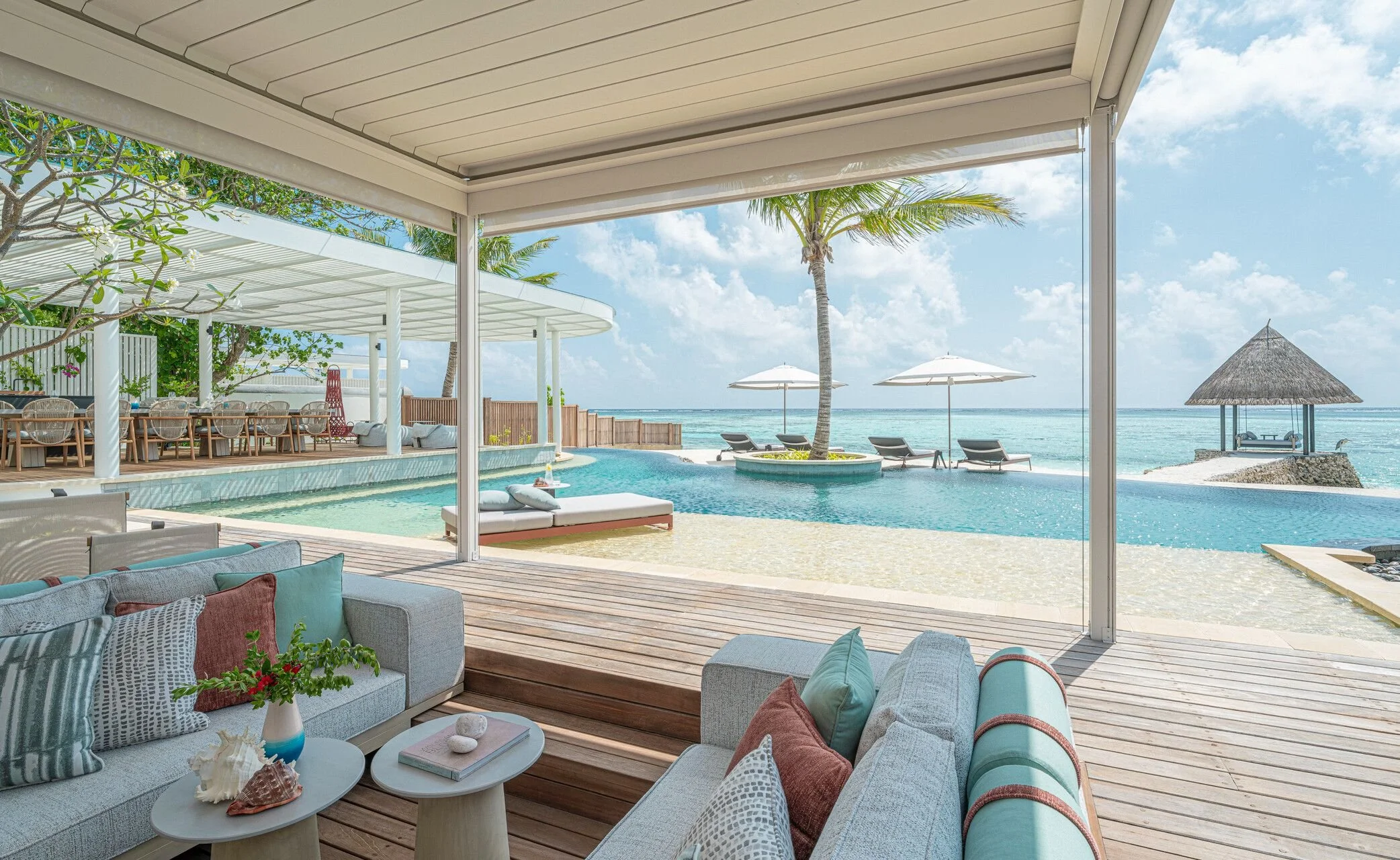 View of a luxurious beachfront resort with a pool, palm trees, and ocean, seen from an open air lounge area with couches and tables.
