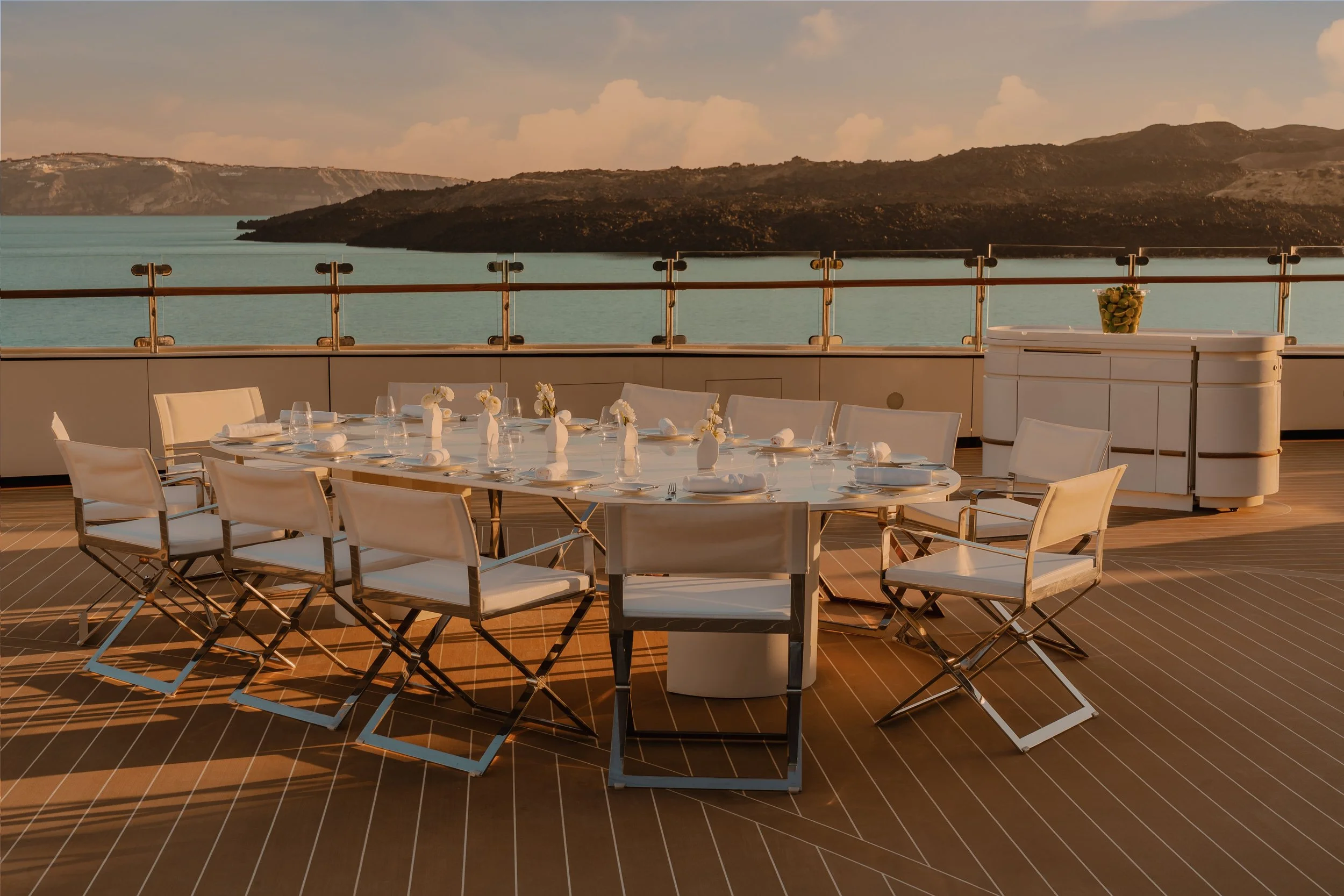 A round dining table set on a balcony with white chairs and white tableware, overlooking a lake with mountains in the distance, during sunset.