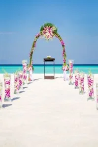 Beach wedding setup with floral arch and seating on white sand, overlooking the ocean under a clear blue sky.