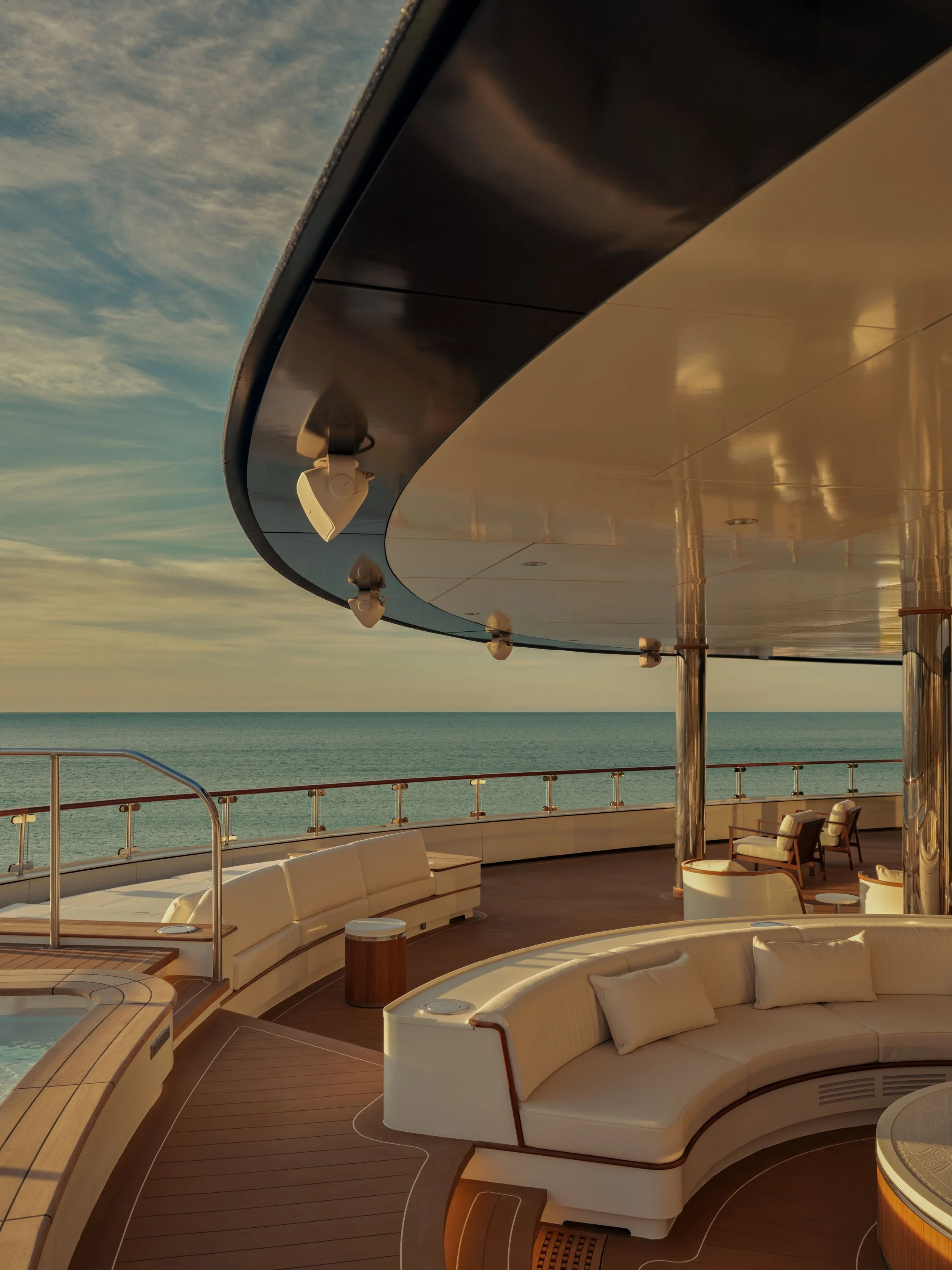 View of the upper deck of a cruise ship with outdoor seating, lounge chairs, and a scenic ocean and sky background during sunset.
