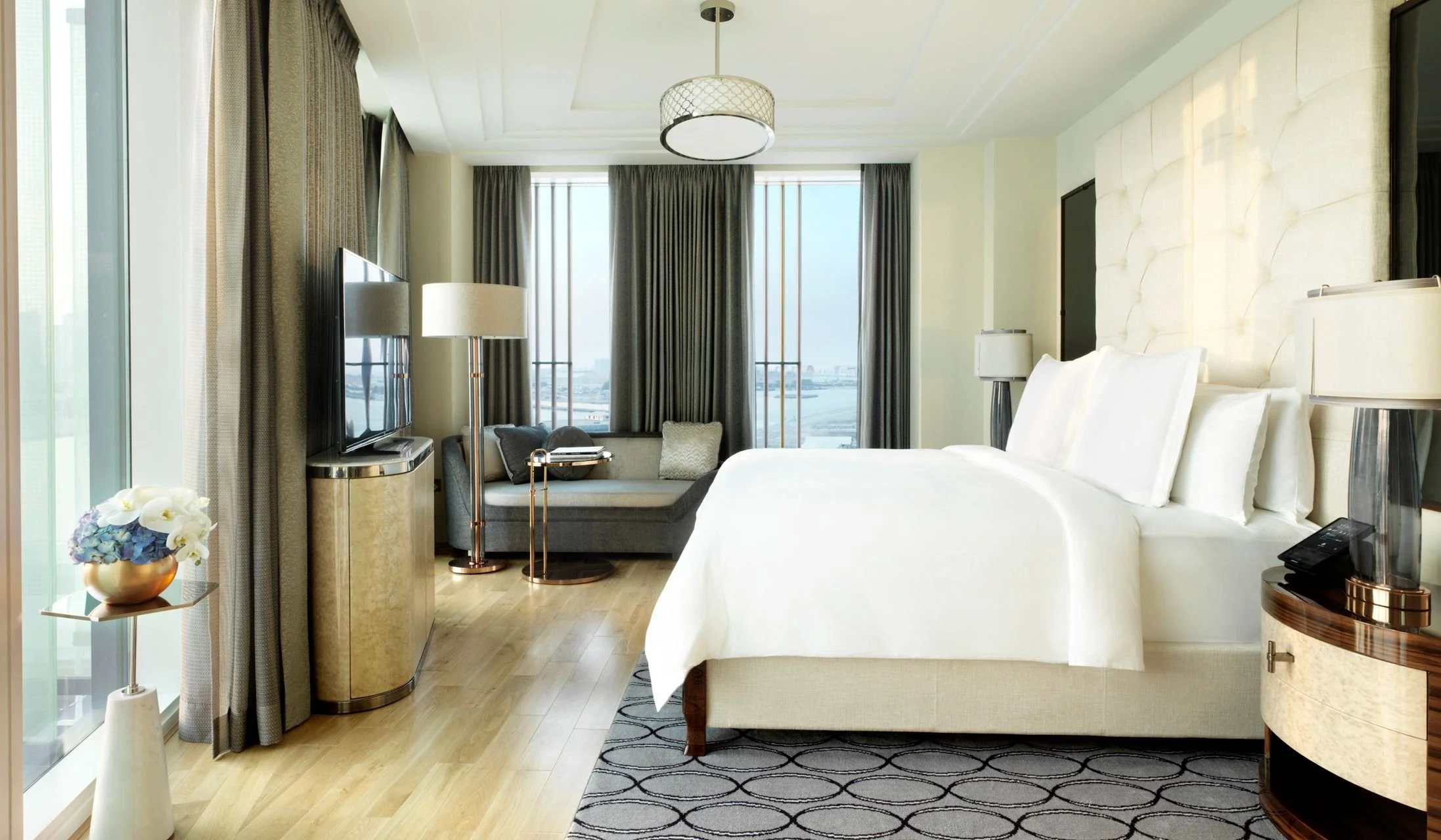 A luxurious hotel room with a large bed, a sitting area with a window seat, a flat-screen TV, and a view of a river or city skyline through large windows, decorated with modern furniture and neutral colors.