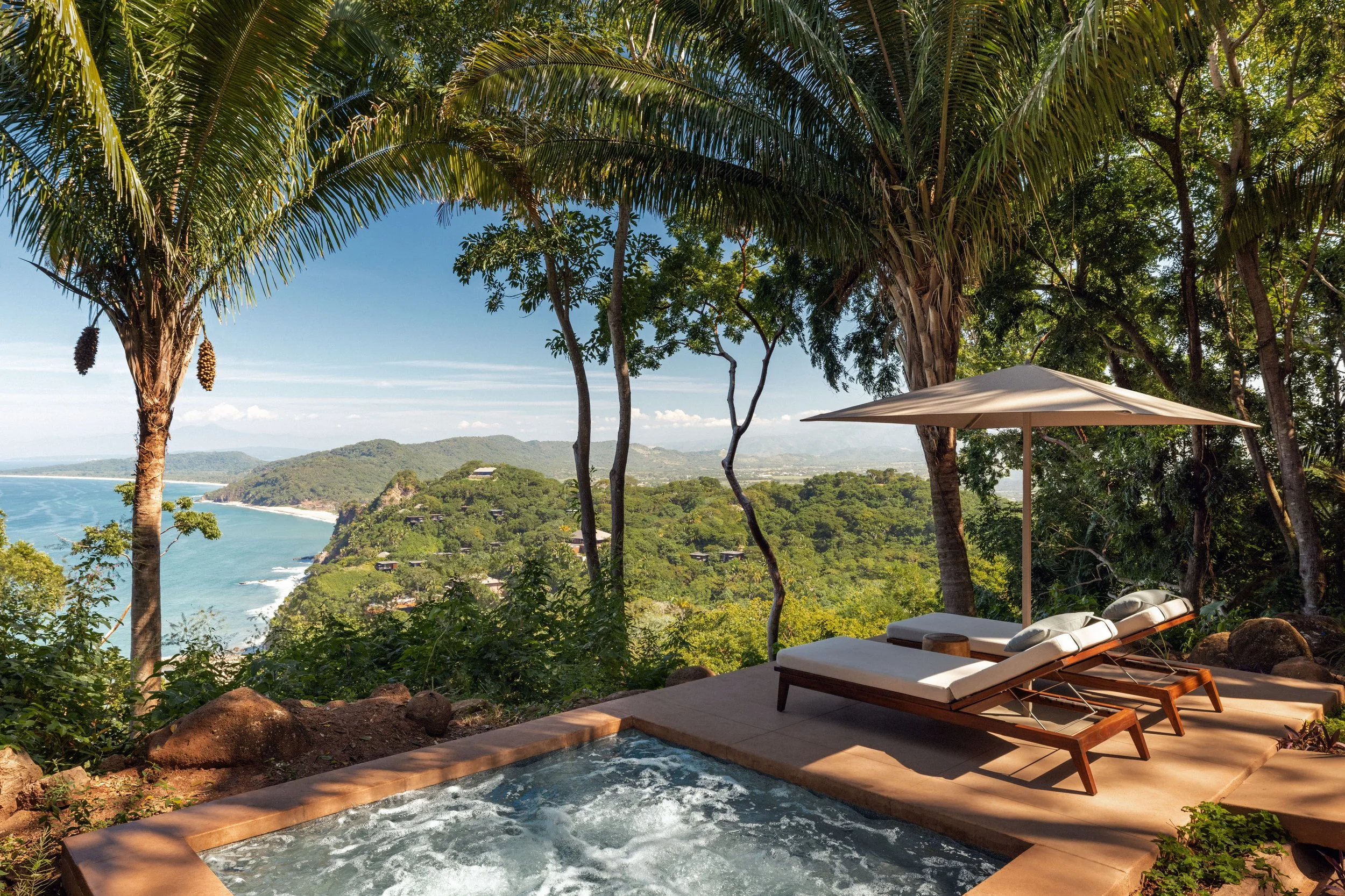 A tropical outdoor scene with lush green trees, a view of the ocean and hills in the background, and a small pool with lounge chairs and an umbrella.