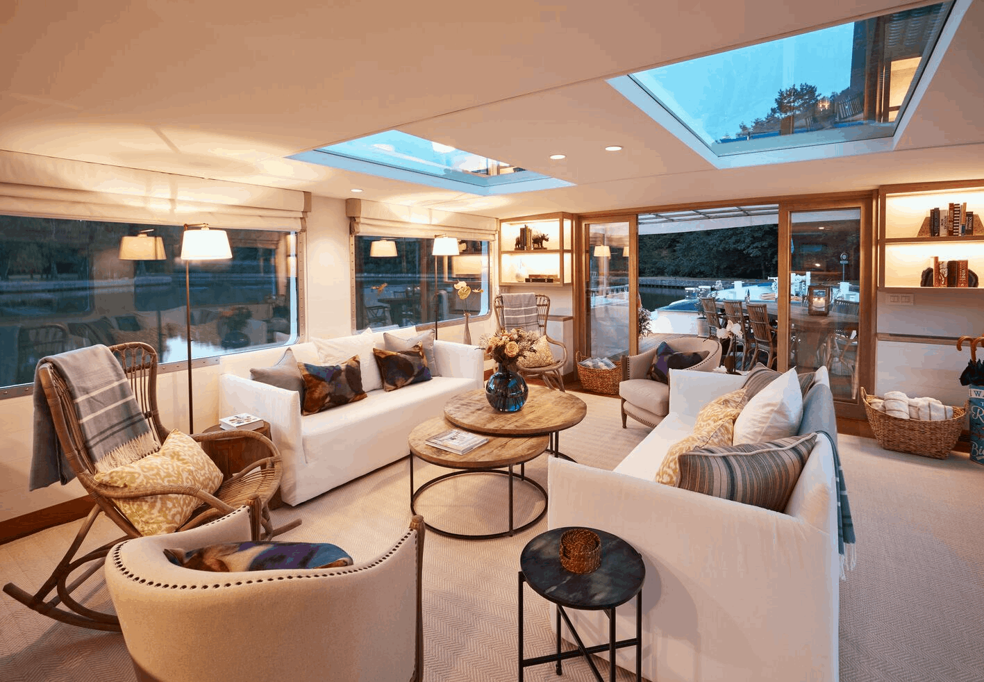 Cozy living room with multiple white sofas, armchairs, wooden tables, and large windows and skylights showing an outdoor patio with wooden chairs and tables.