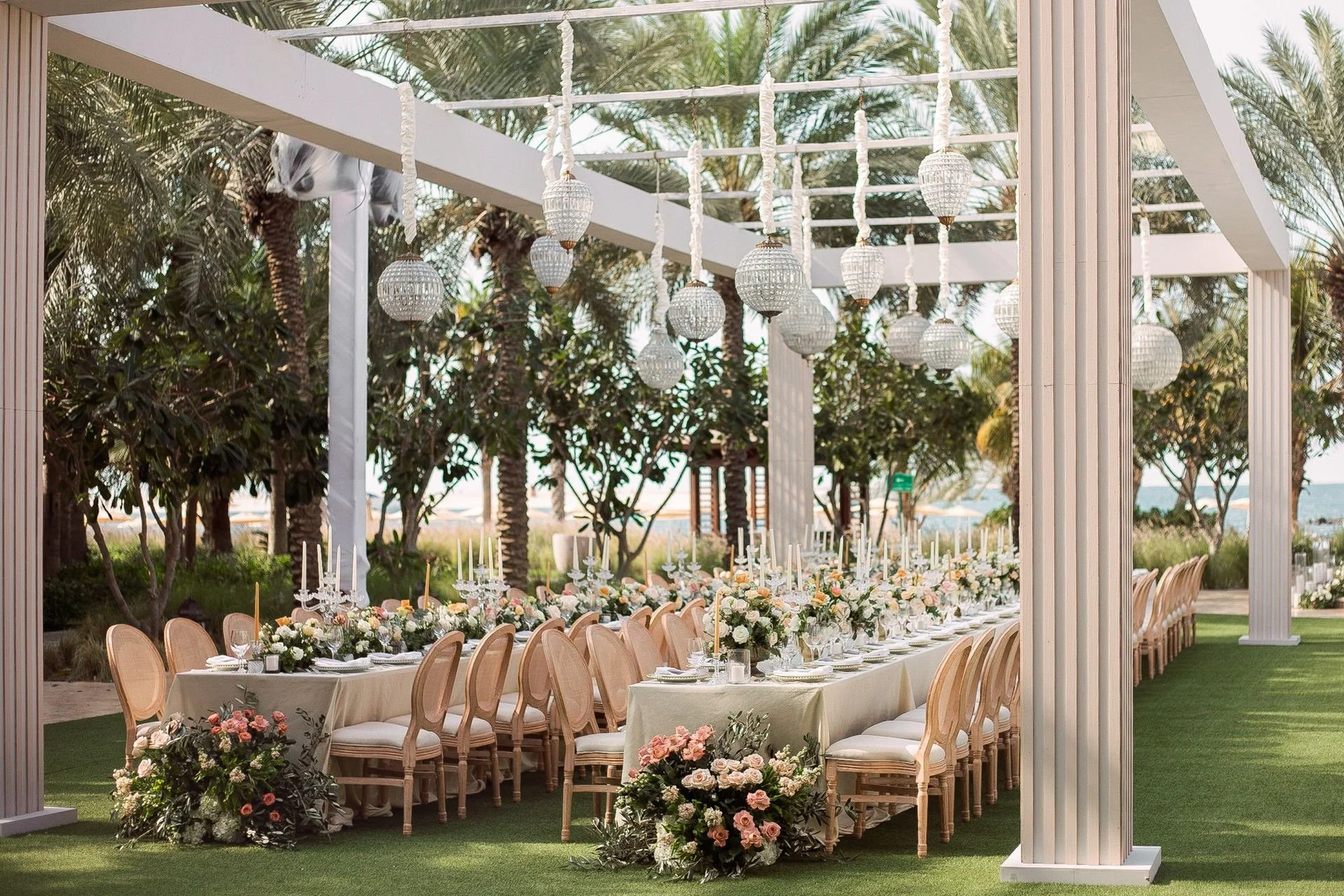 An outdoor wedding or event setup with a long dinner table adorned with white and pastel-colored flowers, surrounded by elegant wooden chairs, under hanging lanterns, with tall palm trees and a beach in the background.