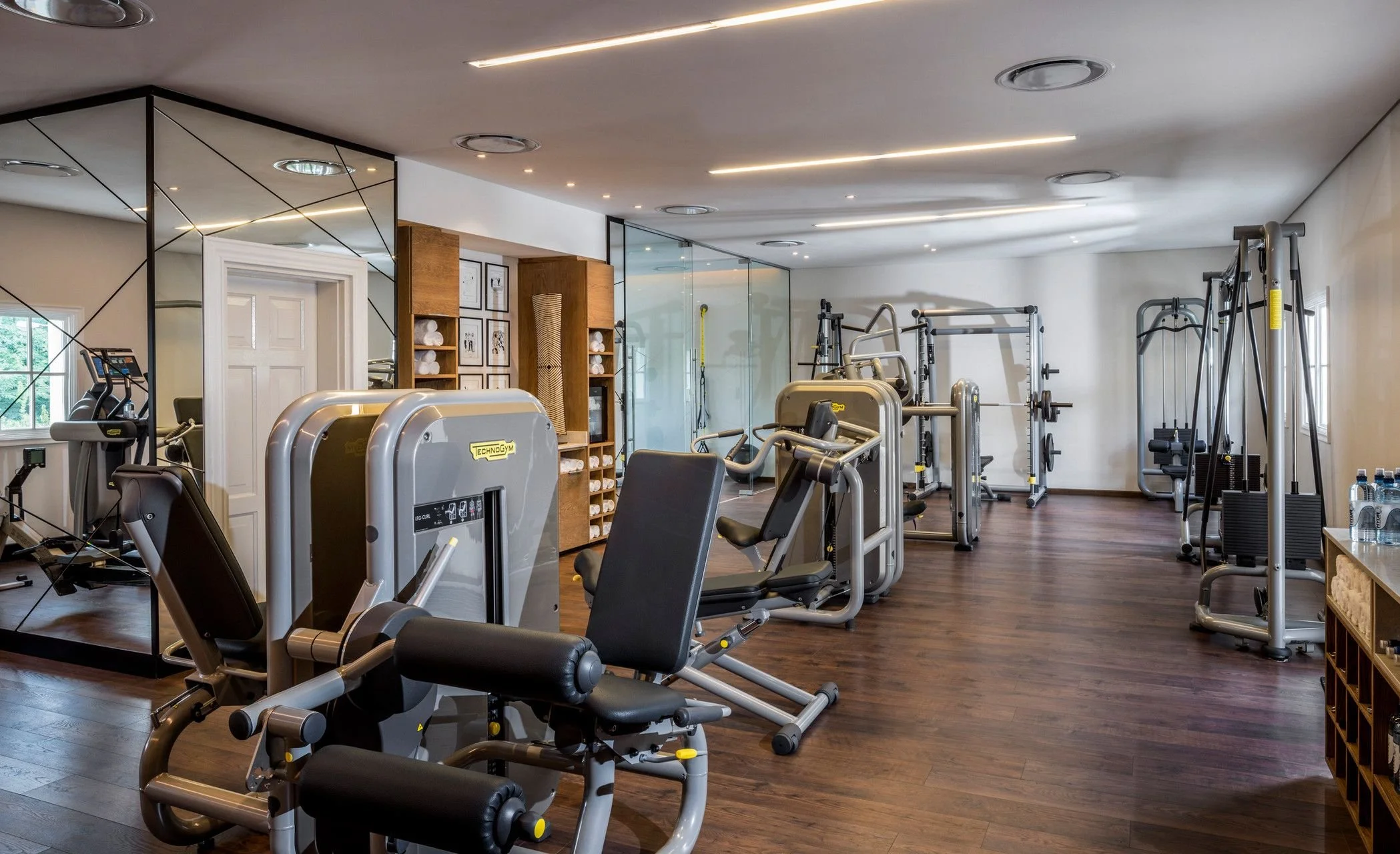 A modern gym with various exercise equipment including treadmills, weight machines, and free weights, with wooden flooring and bright ceiling lighting.