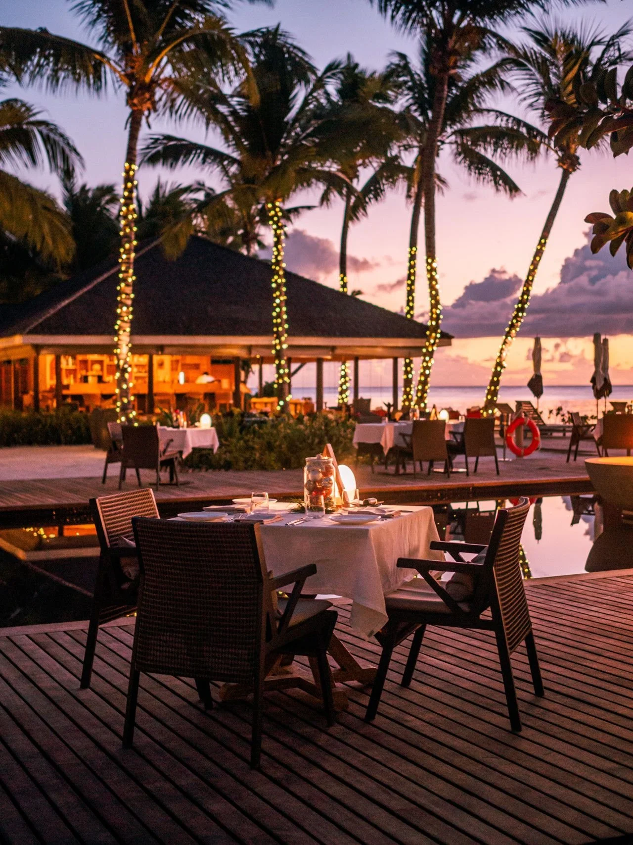 An outdoor dining area on a wooden deck by the beach at sunset, with tables set for dinner and palm trees decorated with string lights.