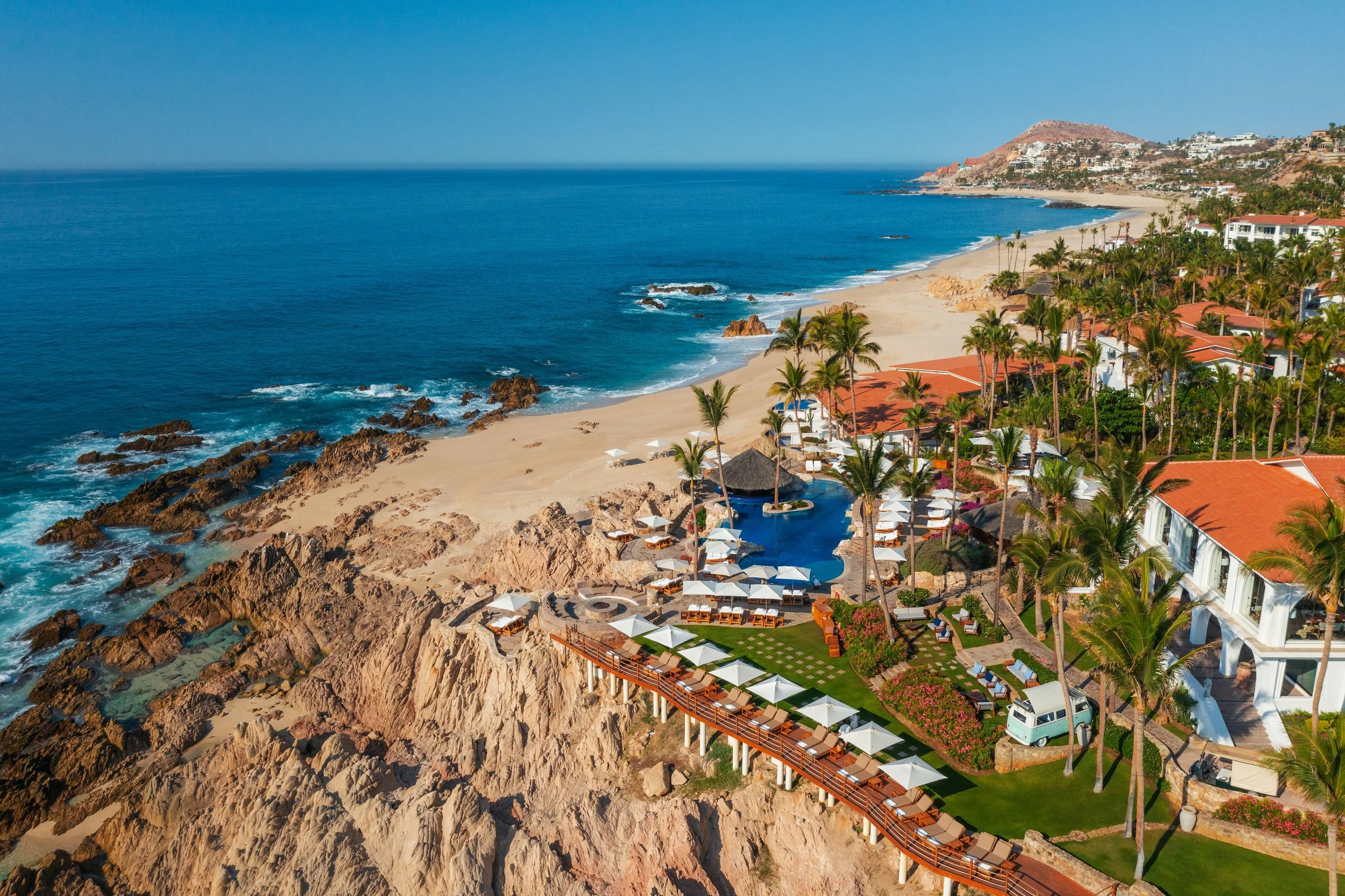 An aerial view of a beachside resort with white buildings, palm trees, lounge chairs, umbrellas, and a beach, with rocky cliffs and the ocean in the background.