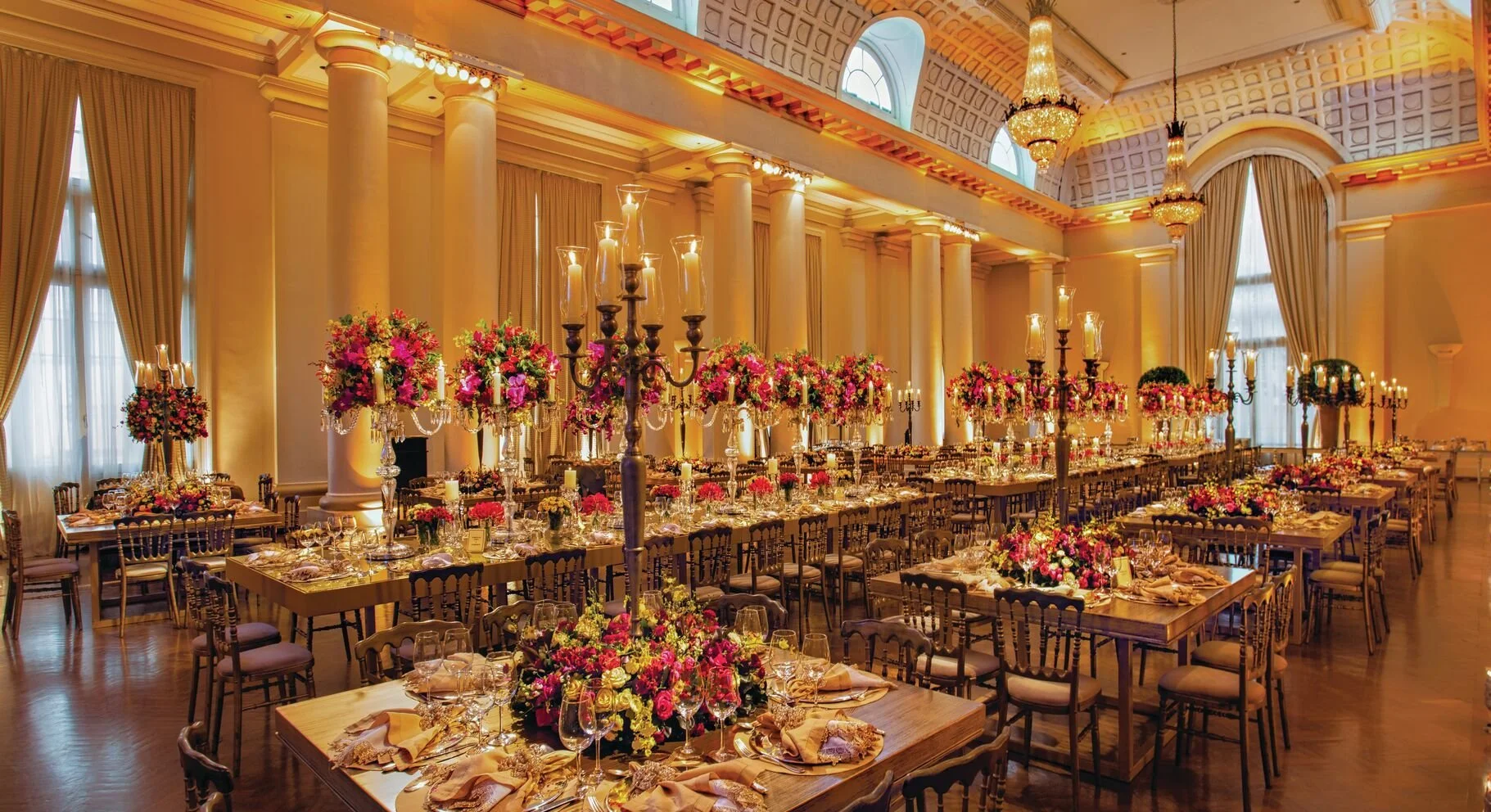 A luxurious banquet hall decorated for a special event with tall floral centerpieces, chandeliers, candelabras, and elegantly set tables with napkins and glassware.