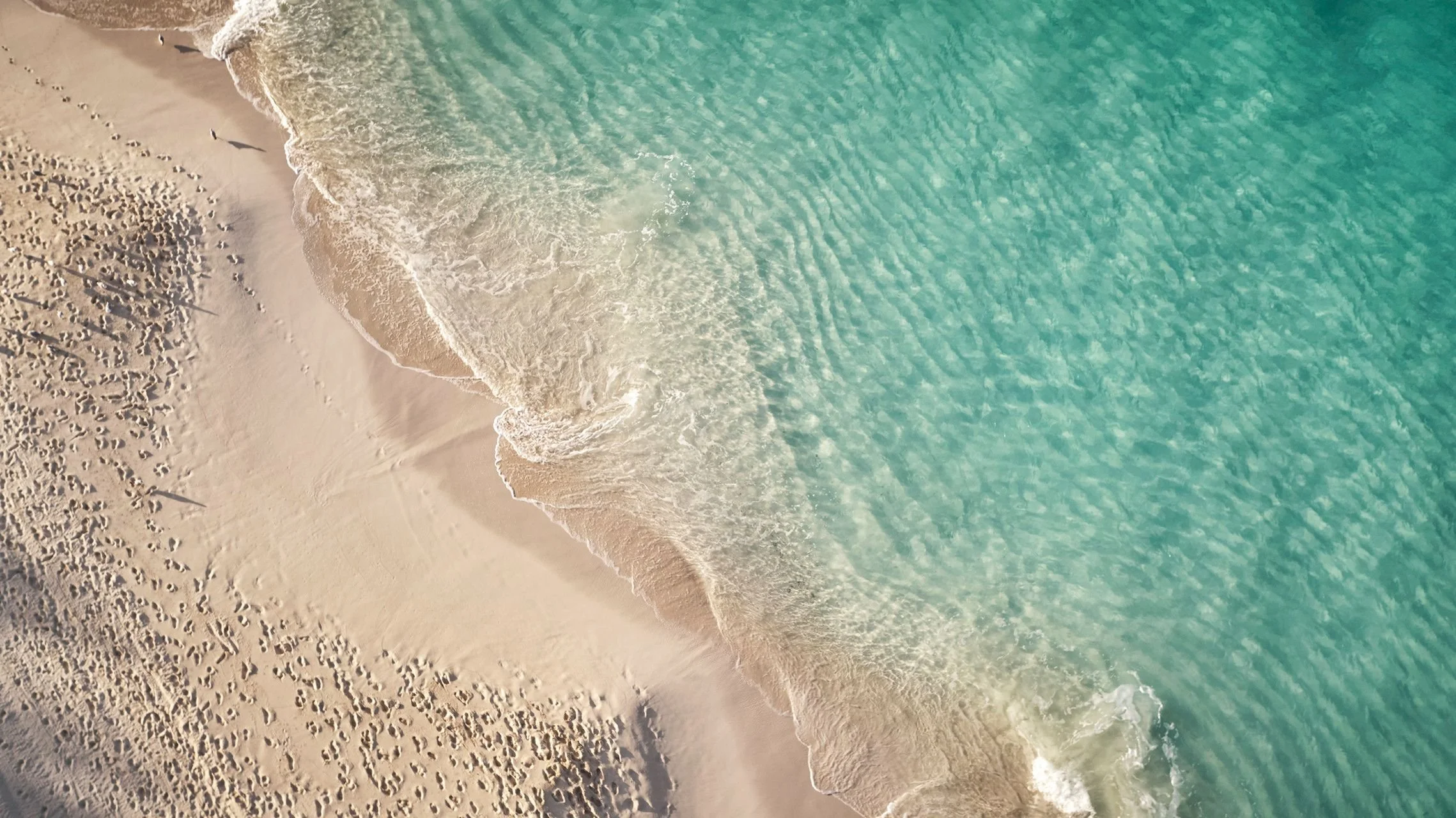 An aerial view of a sandy beach with footprints, ocean waves, and turquoise water.