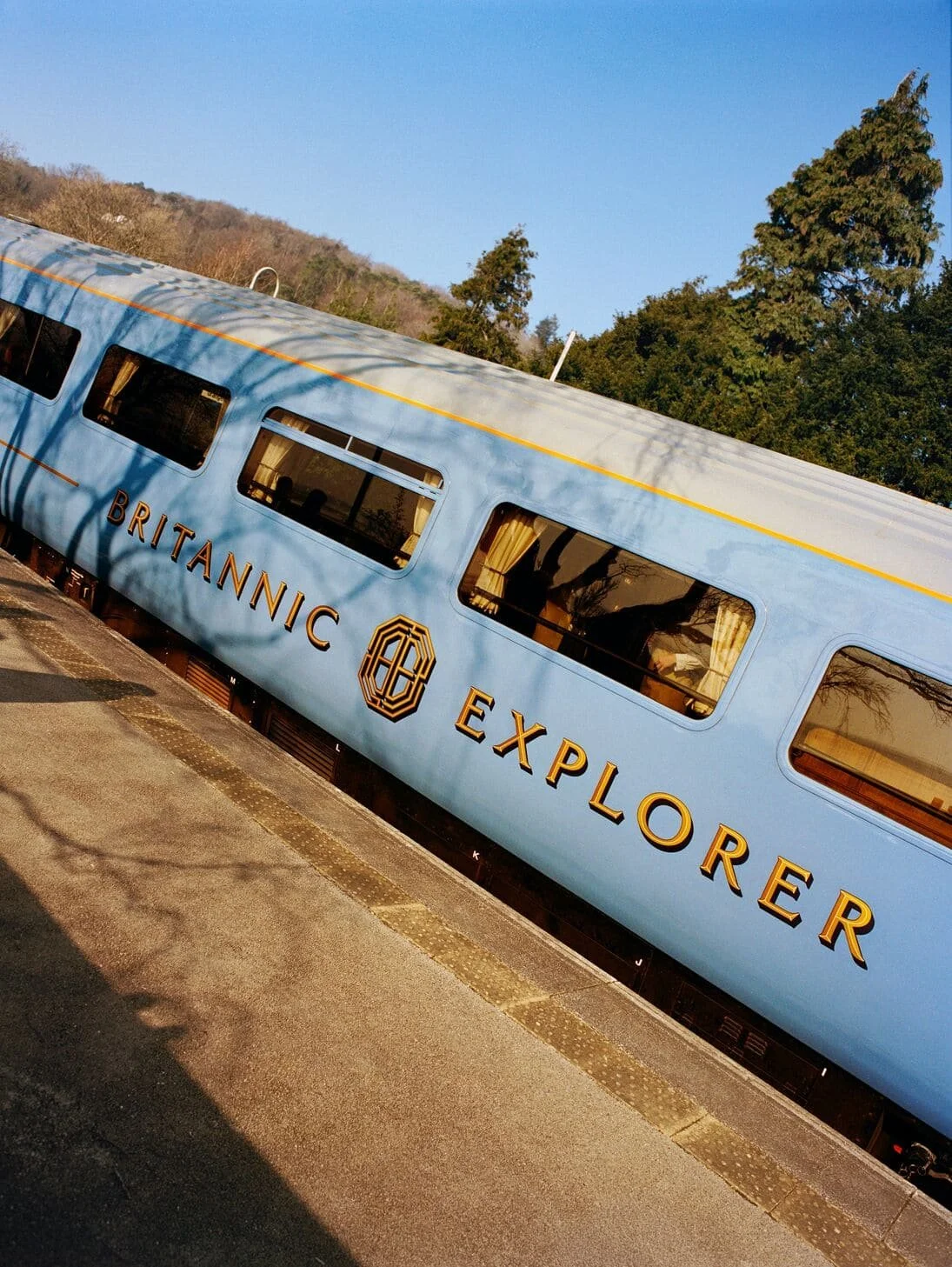 A vintage blue train car with gold lettering that reads 'Britannic Explorer' and a logo, parked at a station with trees and hills in the background.