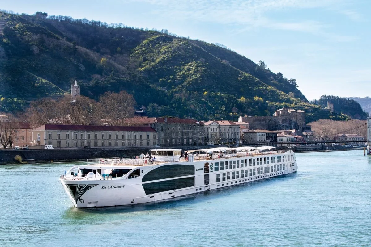 A white cruise ship named S S CATHERINE sailing on a river with a town and green hills in the background.