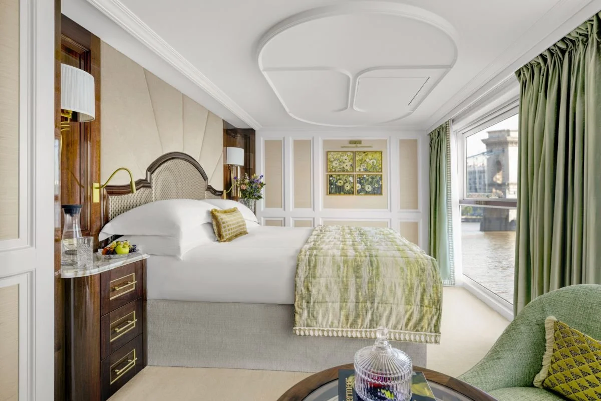A luxurious hotel room with a large bed, green curtains, and artwork of flowers on the wall. A window offers a view of city buildings and water outside.