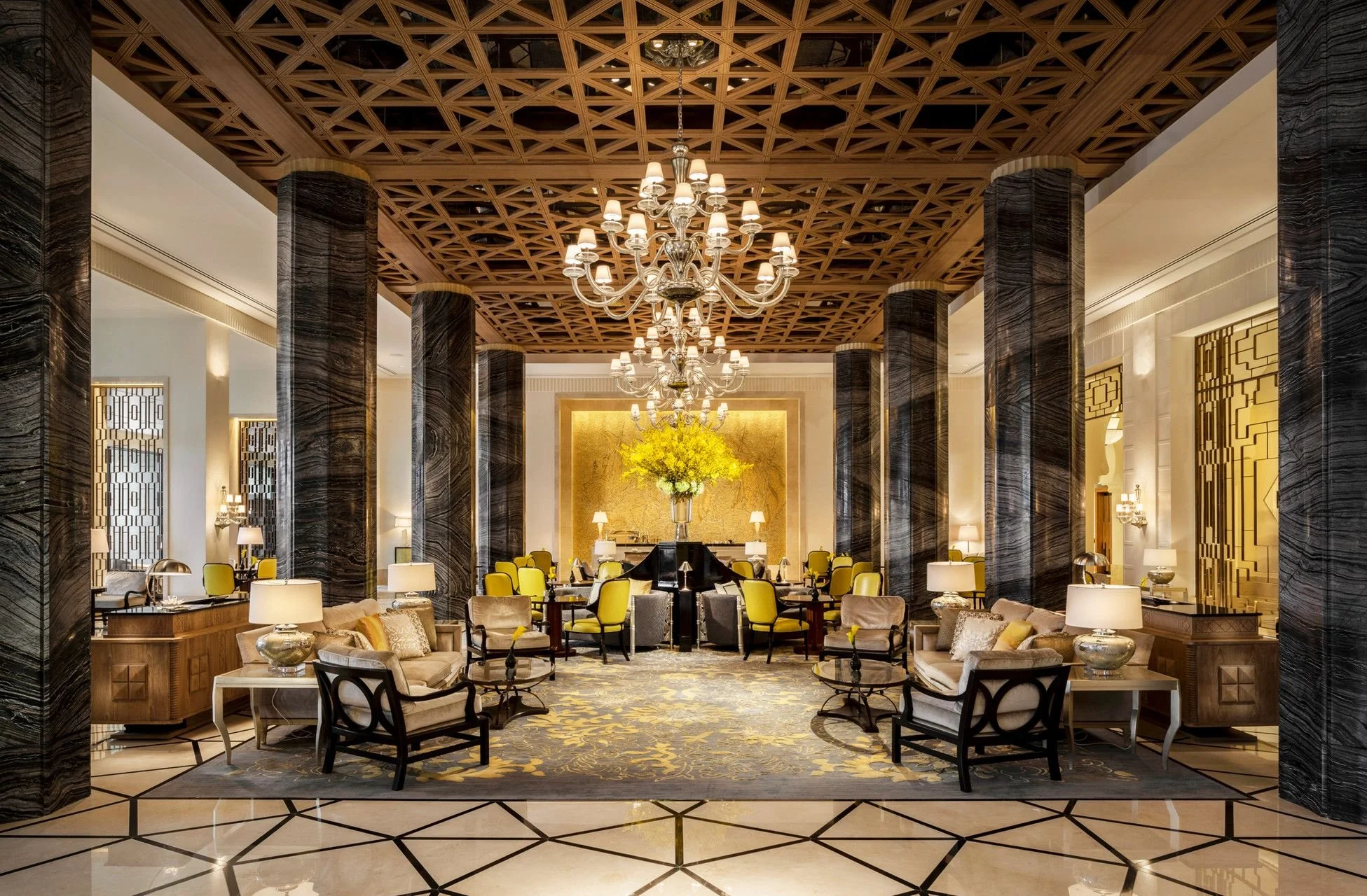 Elegant hotel lobby with grand chandeliers, black marble columns, yellow chairs, and a large floral arrangement with yellow flowers.