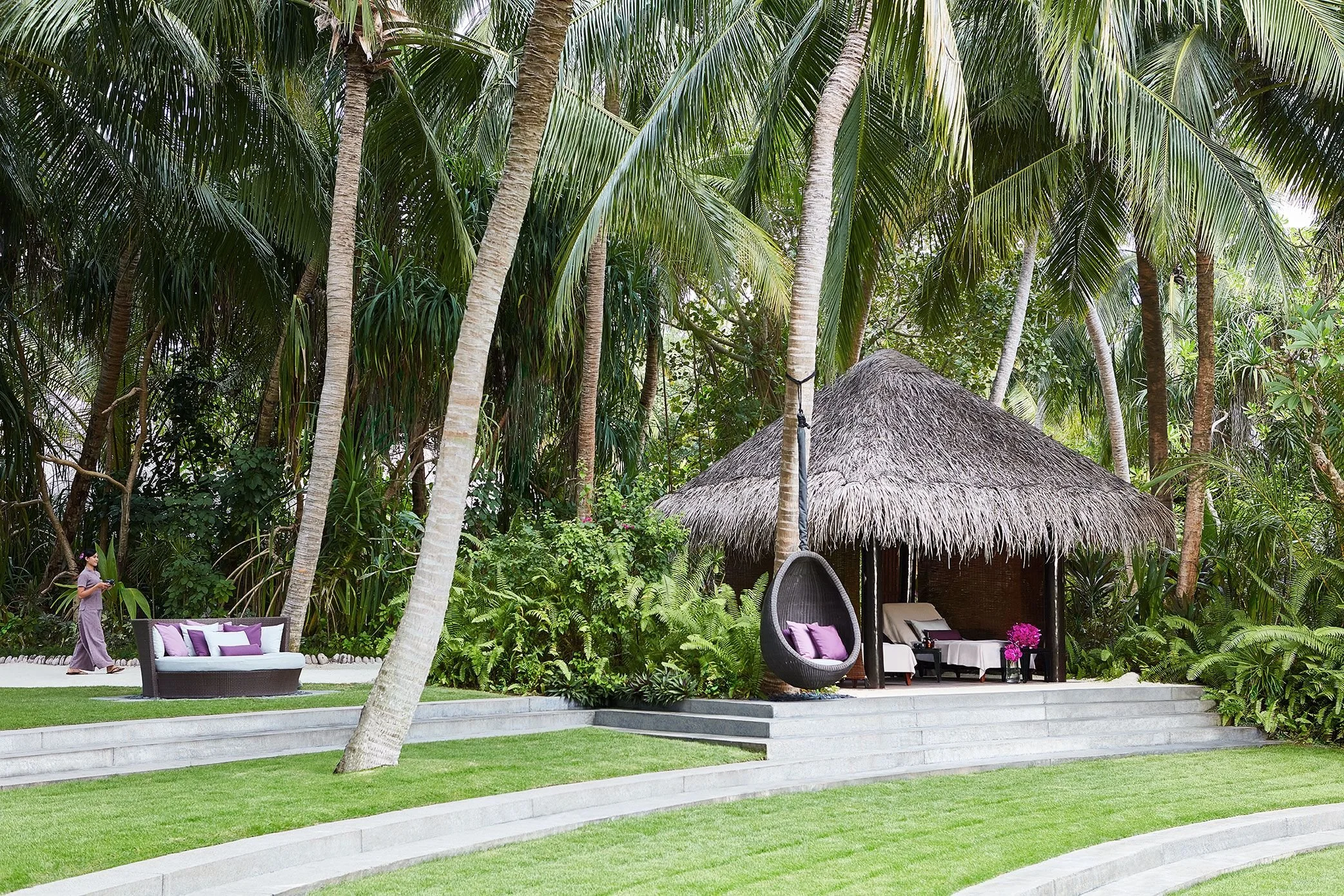 A tropical outdoor setting with tall palm trees, green foliage, a thatched hut, outdoor lounge chairs with purple pillows, and a woman walking nearby.