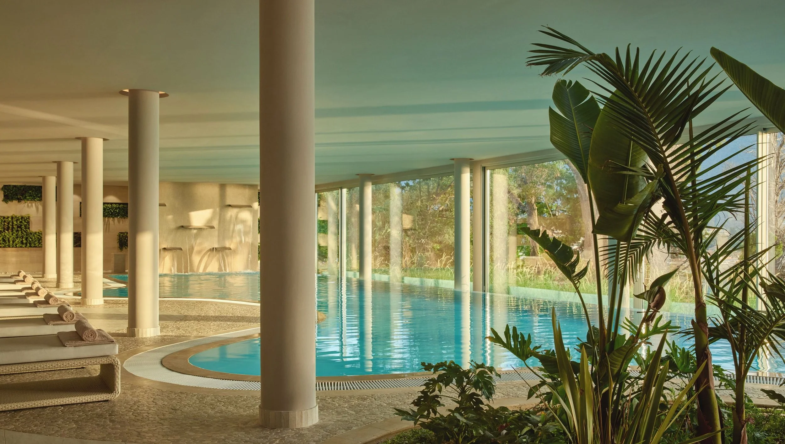 Indoor swimming pool area with lounge chairs, large green plants, and floor-to-ceiling windows showing outdoor trees.
