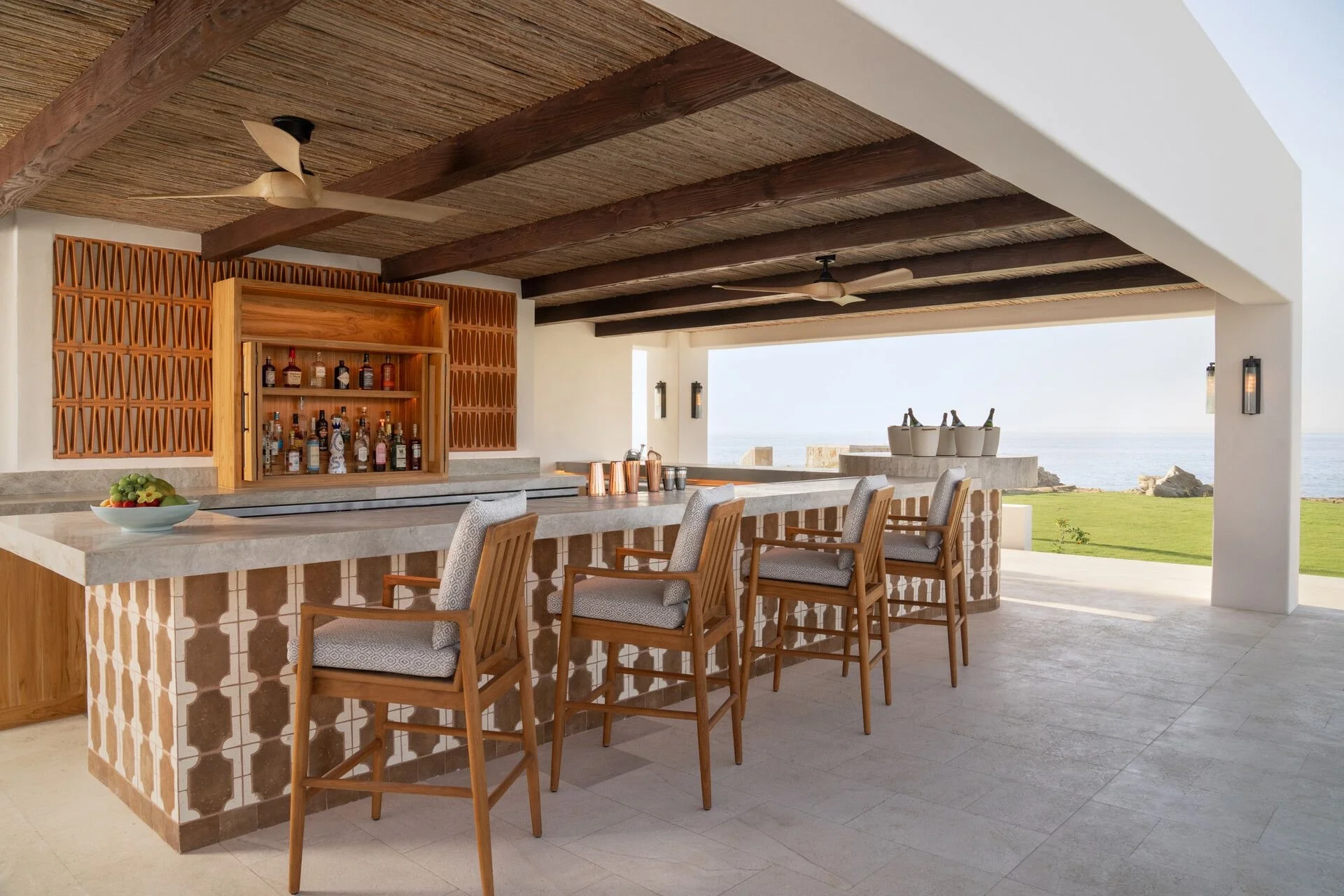 An outdoor bar area with a countertop, bar stools, a wooden cabinet with bottles, and a view of the ocean and grassy area beyond.