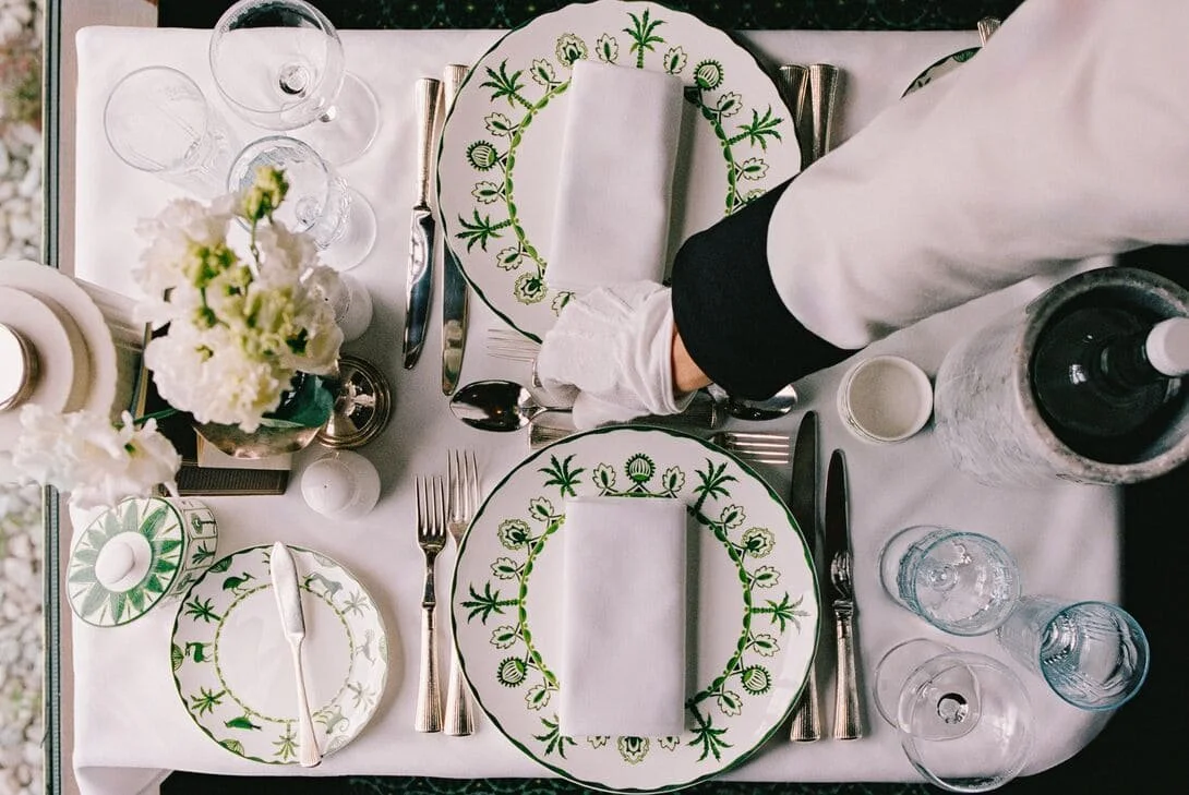 Overhead view of a formal table setting with green and white patterned plates, silverware, glasses, a water bottle, a small white cup, a flower arrangement, and a hand in a white uniform with a black cuff.