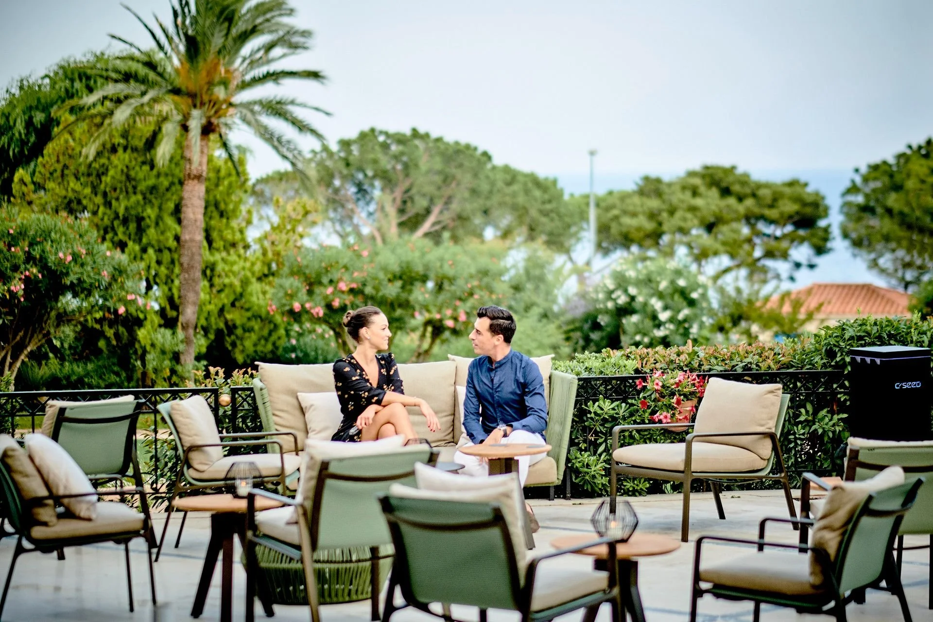 A man and woman sit on a outdoor patio sofa, facing each other and engaging in conversation. The patio is decorated with various chairs and lush greenery, including trees and plants. It appears to be a pleasant, sunny day.