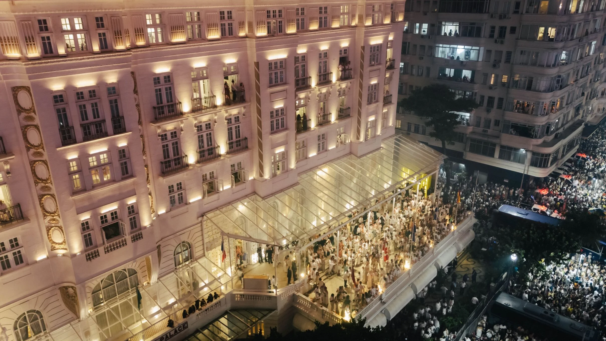Nighttime scene of a large, ornate hotel with many lit windows and balconies. The hotel is filled with a crowd of people on the balconies and on the street below, with some visible umbrellas and flags.