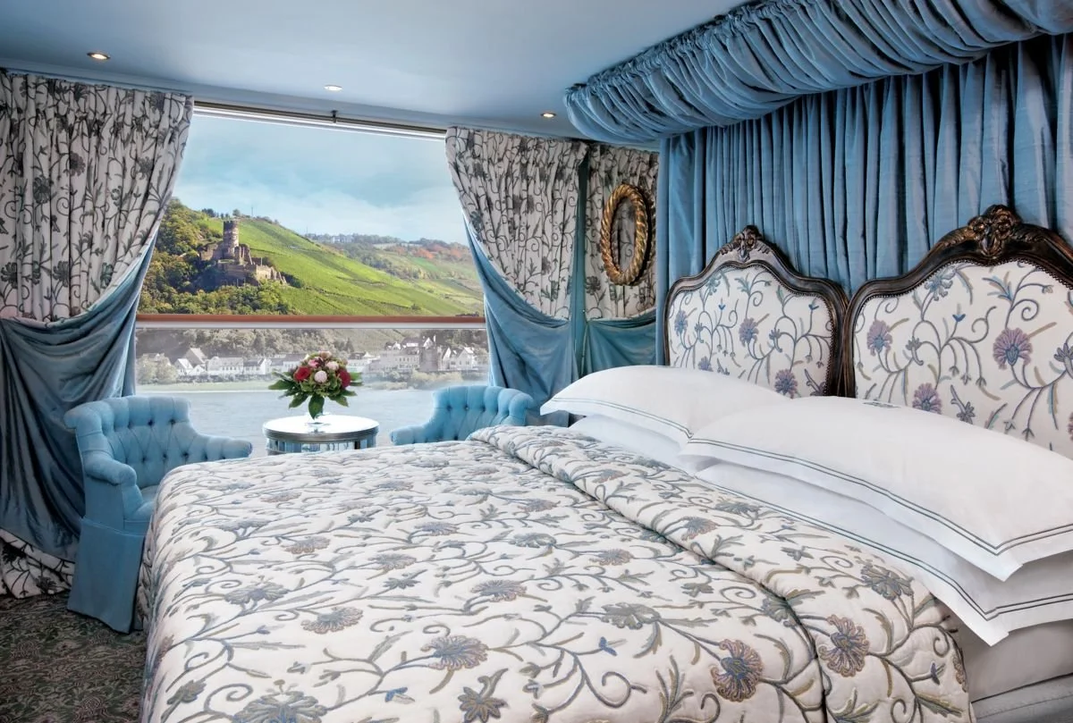 Luxurious hotel room with a large bed, floral patterned bedding, upholstered headboard, and curtains. A window shows a scenic view of green hills with a castle ruin on top and a small town below.