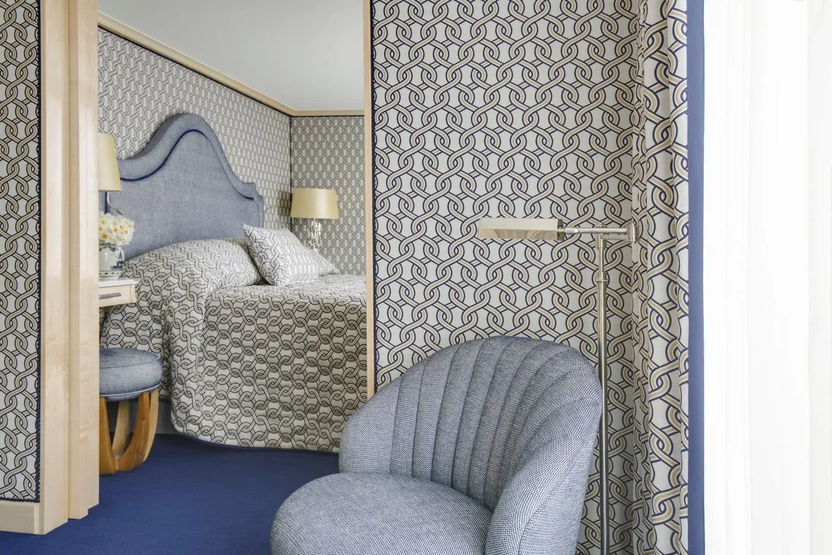 A cozy bedroom with walls covered in a geometric patterned wallpaper. A bed with a blue upholstered headboard and matching bedspread and pillows. A bedside table with a lamp and a vase of flowers. In the foreground, a rounded gray upholstered chair and a standing lamp with an adjustable arm near a window with white curtains.