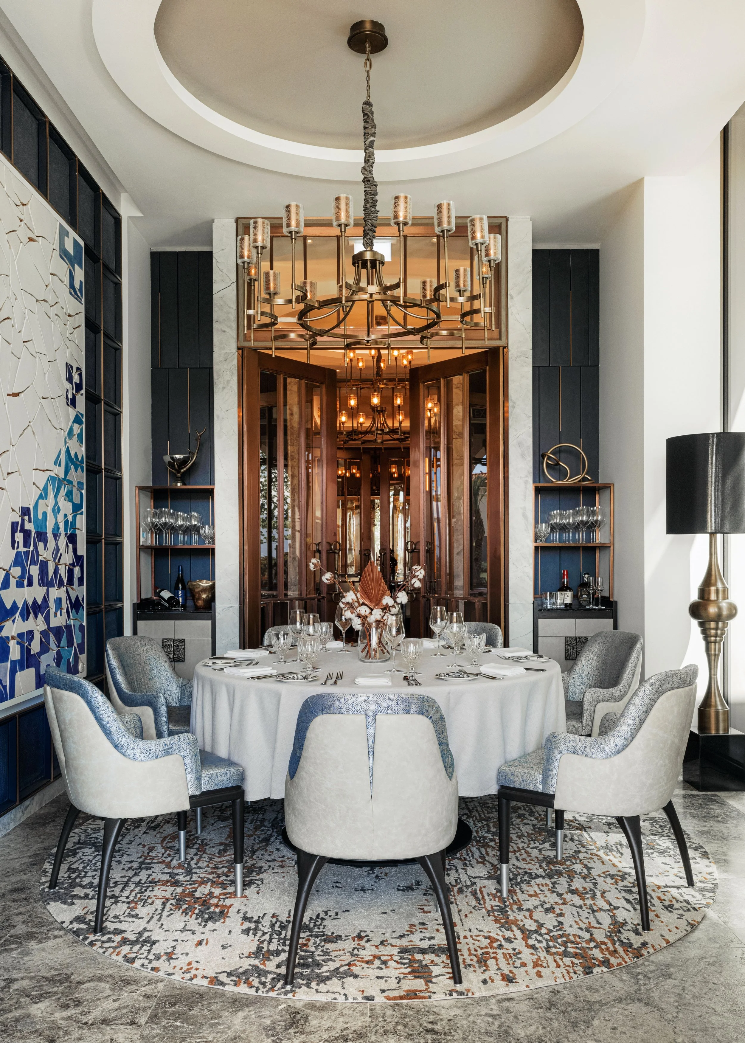 Elegant dining room with a round table, white tablecloth, and 8 chairs in light upholstery. Set for a meal with glassware, silverware, and a floral centerpiece. Chandeliers hang above, reflected in large mirrors behind. Decorative shelves with wine and glassware on either side. Marble flooring and a patterned rug.