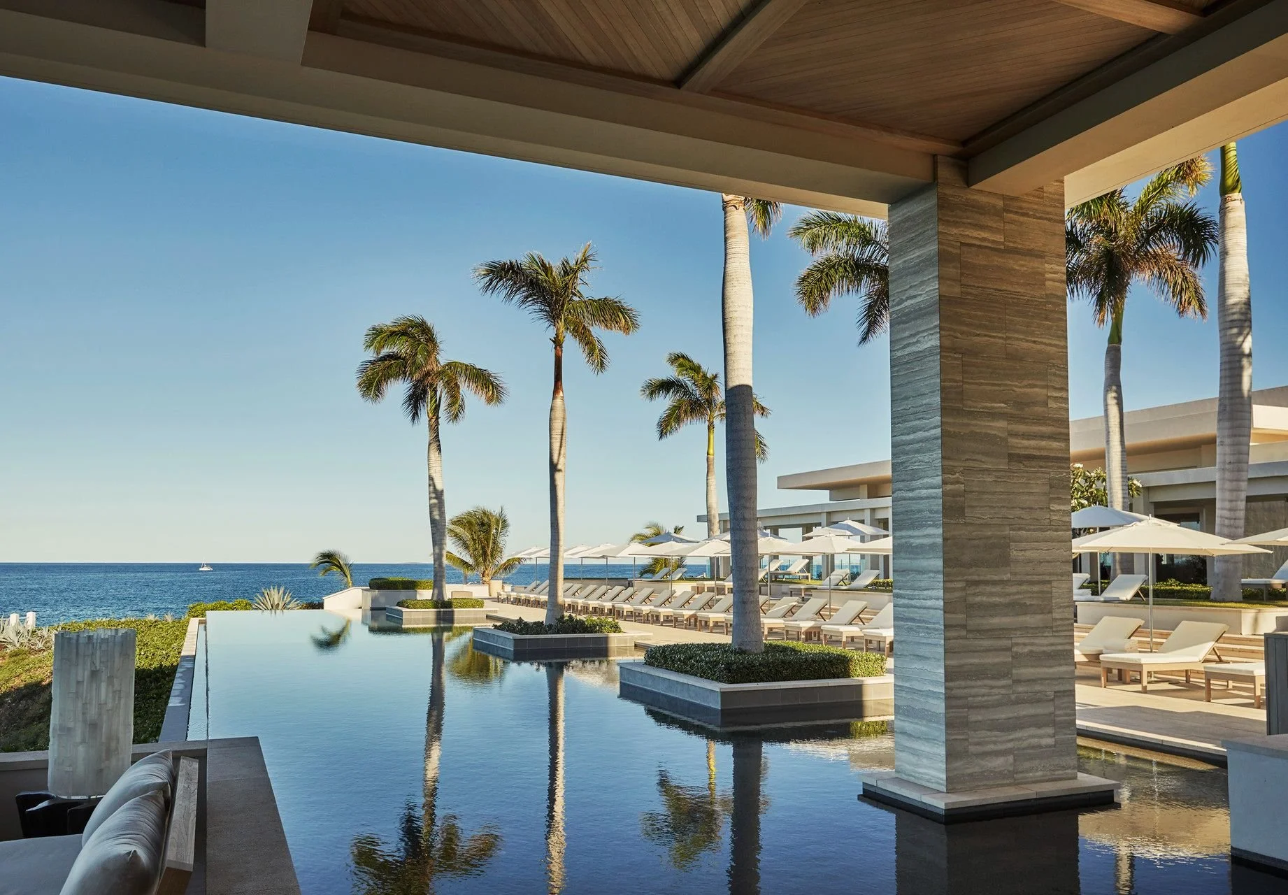 View of a luxurious outdoor pool area with lounge chairs, umbrellas, and palm trees overlooking the ocean under a blue sky.