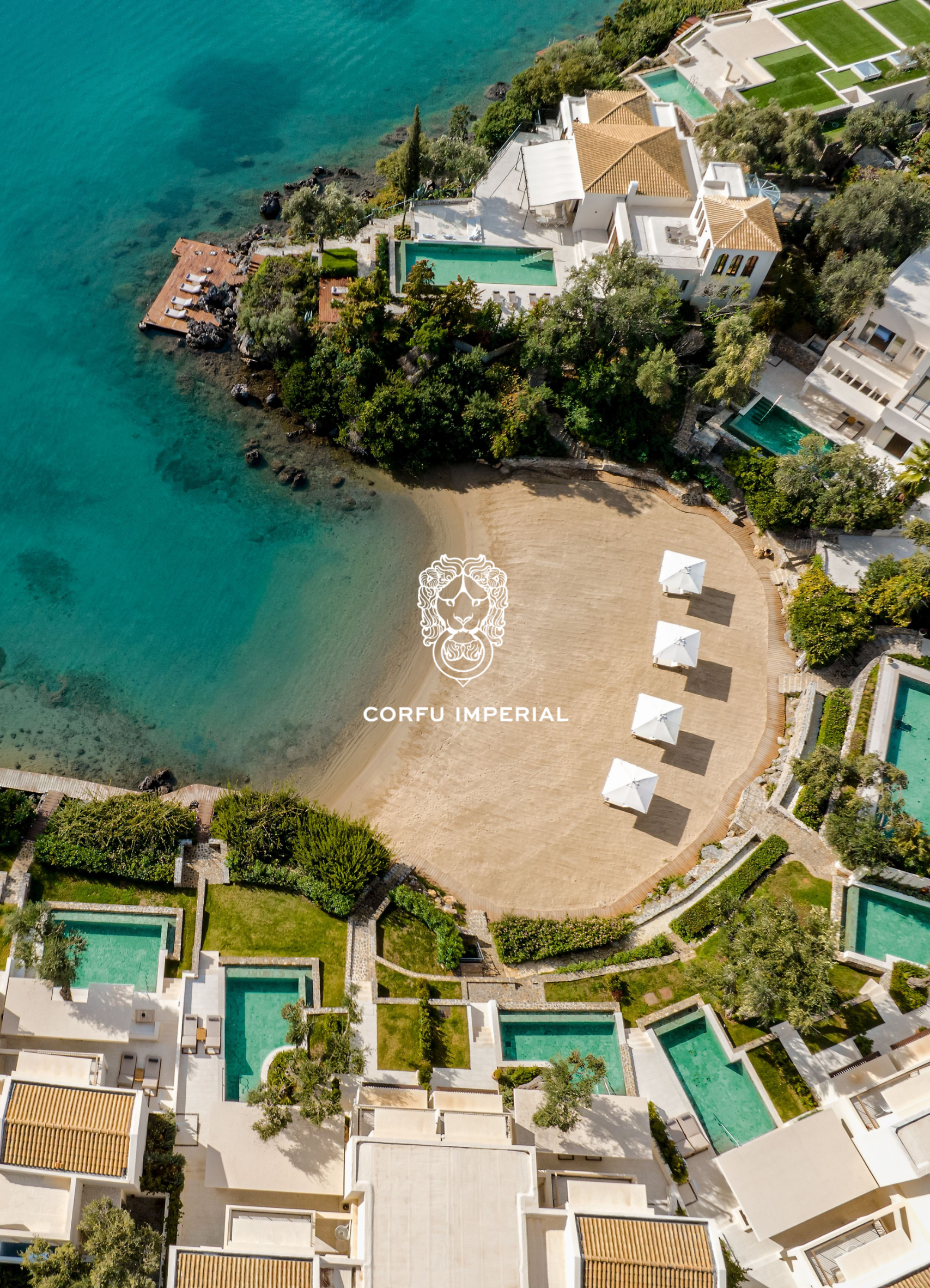 Aerial view of a coastal resort with multiple white buildings, swimming pools, and a beach with umbrellas. The village is situated along the shoreline with turquoise water and rocky coastline. Green trees and landscaped areas are also visible.