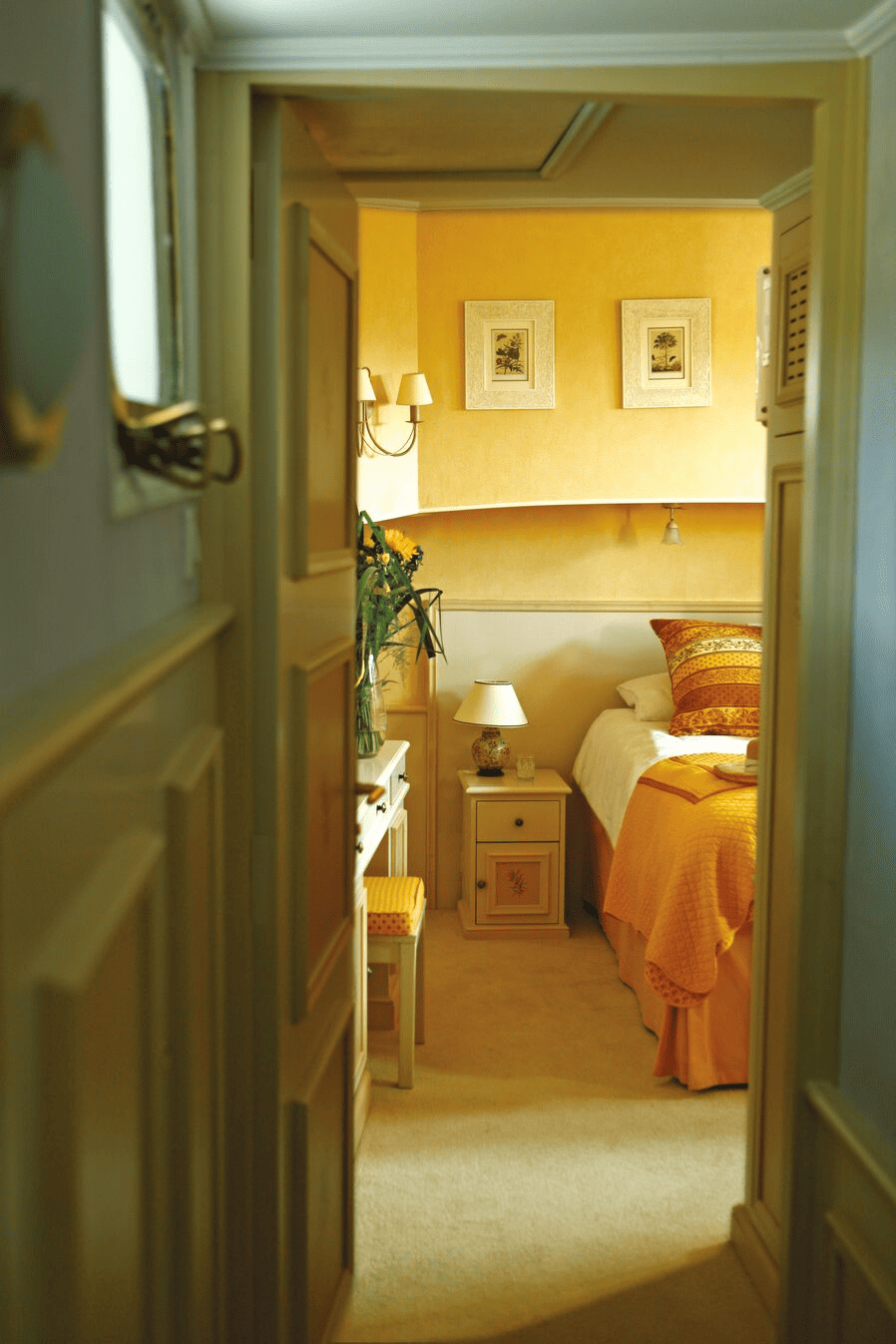 View into a cozy bedroom with a bed, colorful bedding, bedside table with a lamp, and framed artwork on the wall, seen through an open doorway.