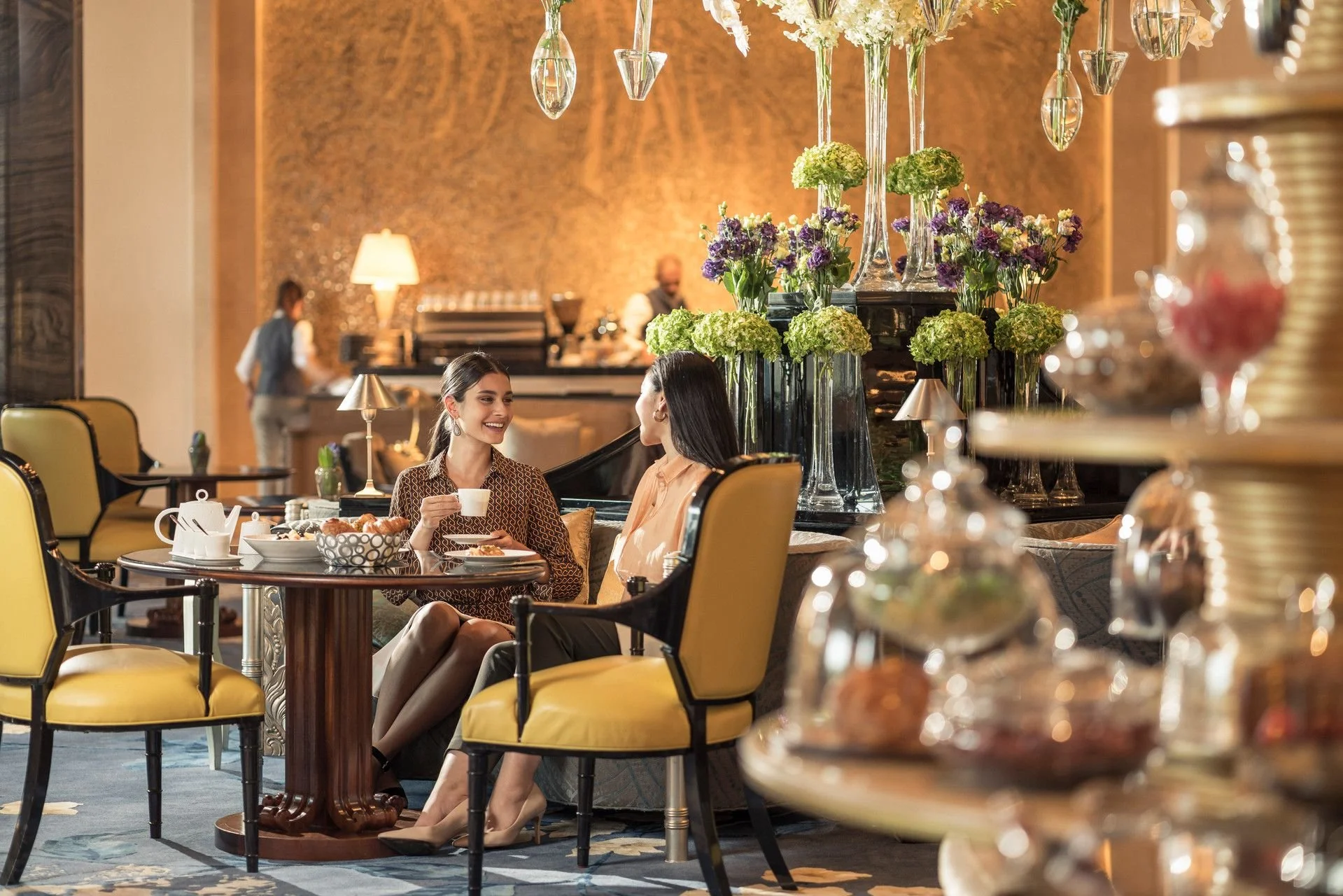 Two women are sitting at a table in a luxurious hotel lounge or restaurant, smiling and having a conversation. The table has a teapot, cups, and snacks. Large floral arrangements with green and purple flowers and hanging glass decor decorate the background. The atmosphere is warm and elegant.