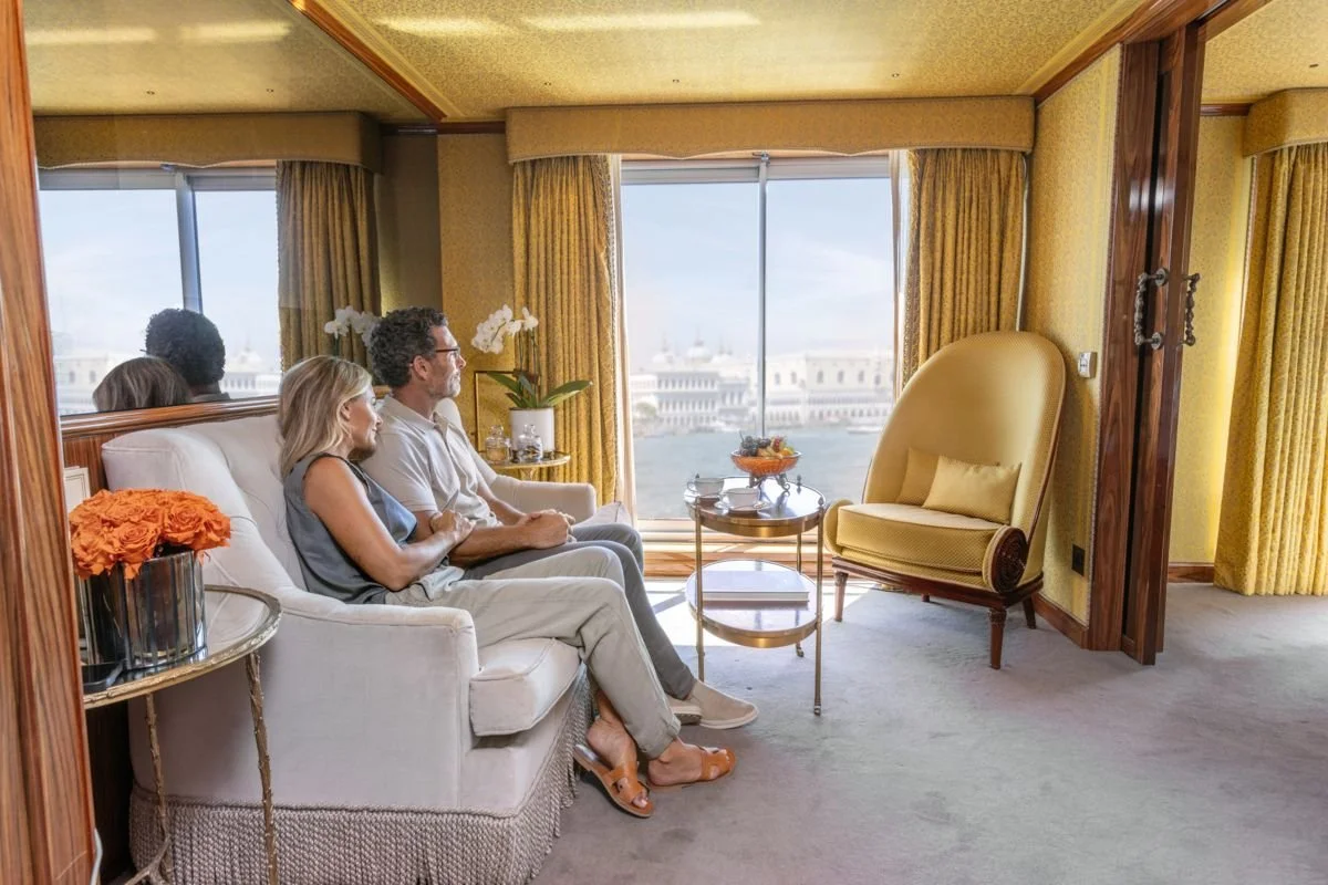 A couple sitting on a sofa in a hotel room, looking out at a harbor view through large windows with yellow curtains. The room is decorated with yellow and gold tones, with a side table holding flowers and a glass bowl of fruit, and a yellow armchair nearby.