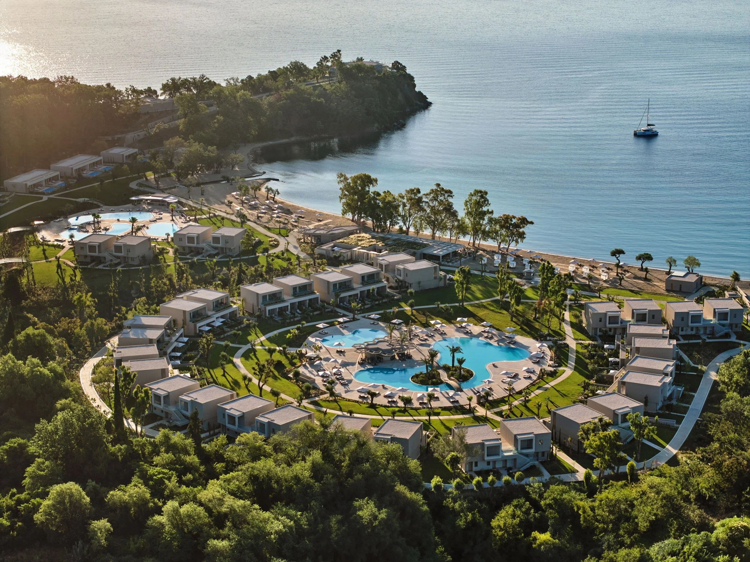 Aerial view of a coastal resort with pools, white modern buildings, green trees, and a beach with umbrellas, overlooking the ocean with a sailboat.