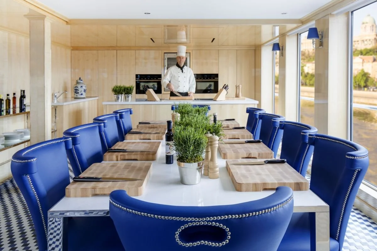 A chef in a white uniform and hat stands in a modern restaurant kitchen behind a table, which is set with cooking utensils, and wood cutting boards. The dining area has a white table with a row of potted herbs, pepper and salt shakers, wine bottles, and blue upholstered chairs around it. Large windows with blue wall sconces provide a view outside of a cityscape and river.