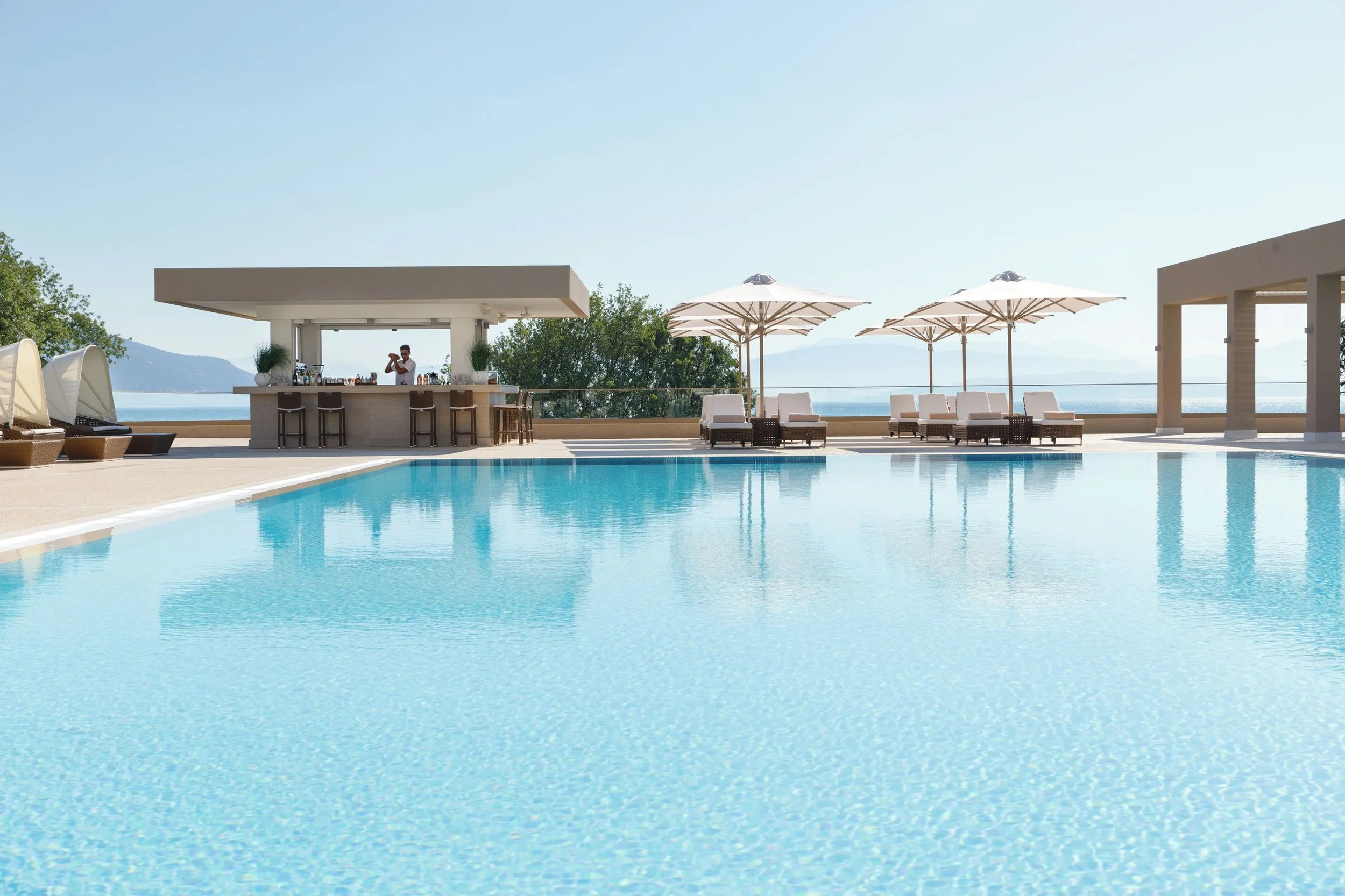 Luxury outdoor pool area with lounge chairs, umbrellas, and a bar by the water, overlooking distant mountains under a clear blue sky.