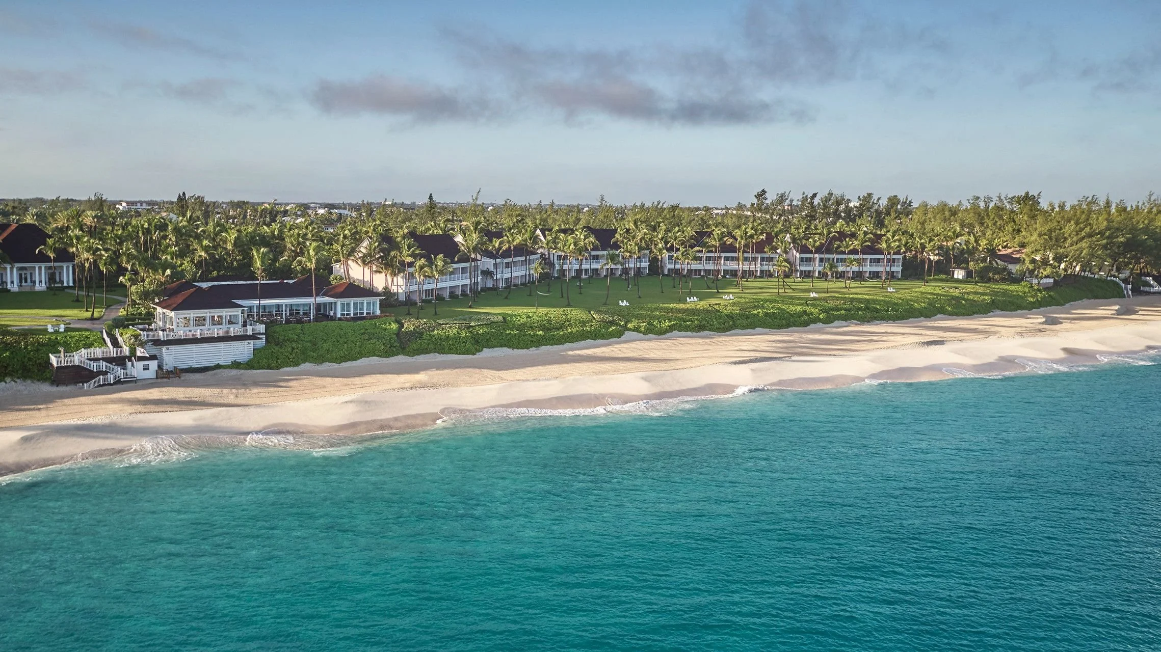 Aerial view of beachfront resort with white buildings, palm trees, green lawns, sandy beach, and turquoise ocean waters under cloudy sky.