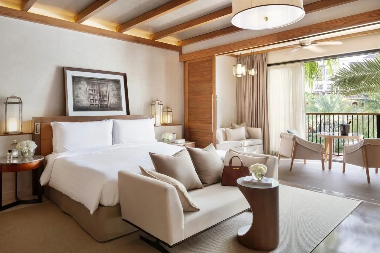 A luxurious hotel room with a large bed, cozy seating area, and balcony view of palm trees and greenery.