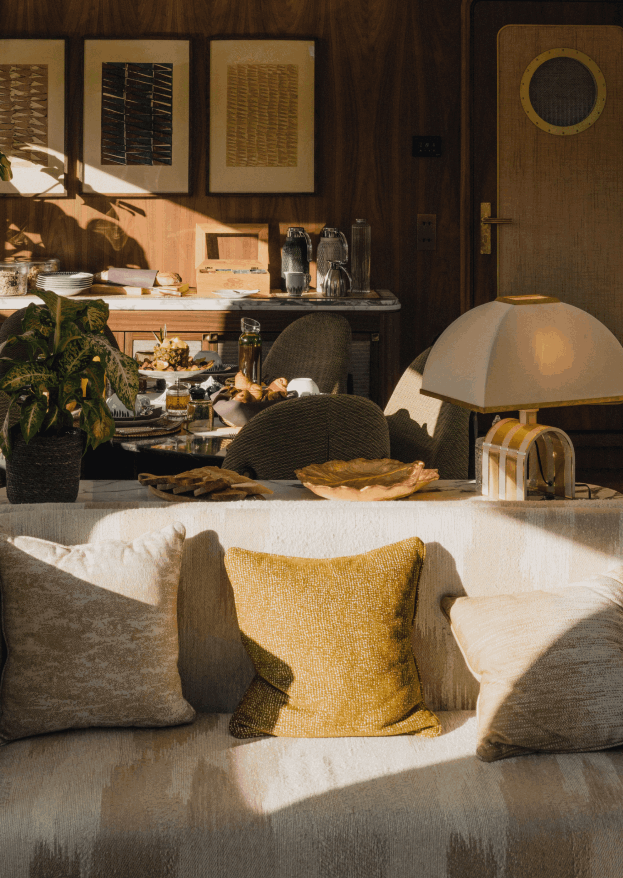 Living room with a beige sofa, a mustard throw pillow, a potted plant, and a dining table set with dishes and a cake. There's a wooden wall with paintings and a door on the right.