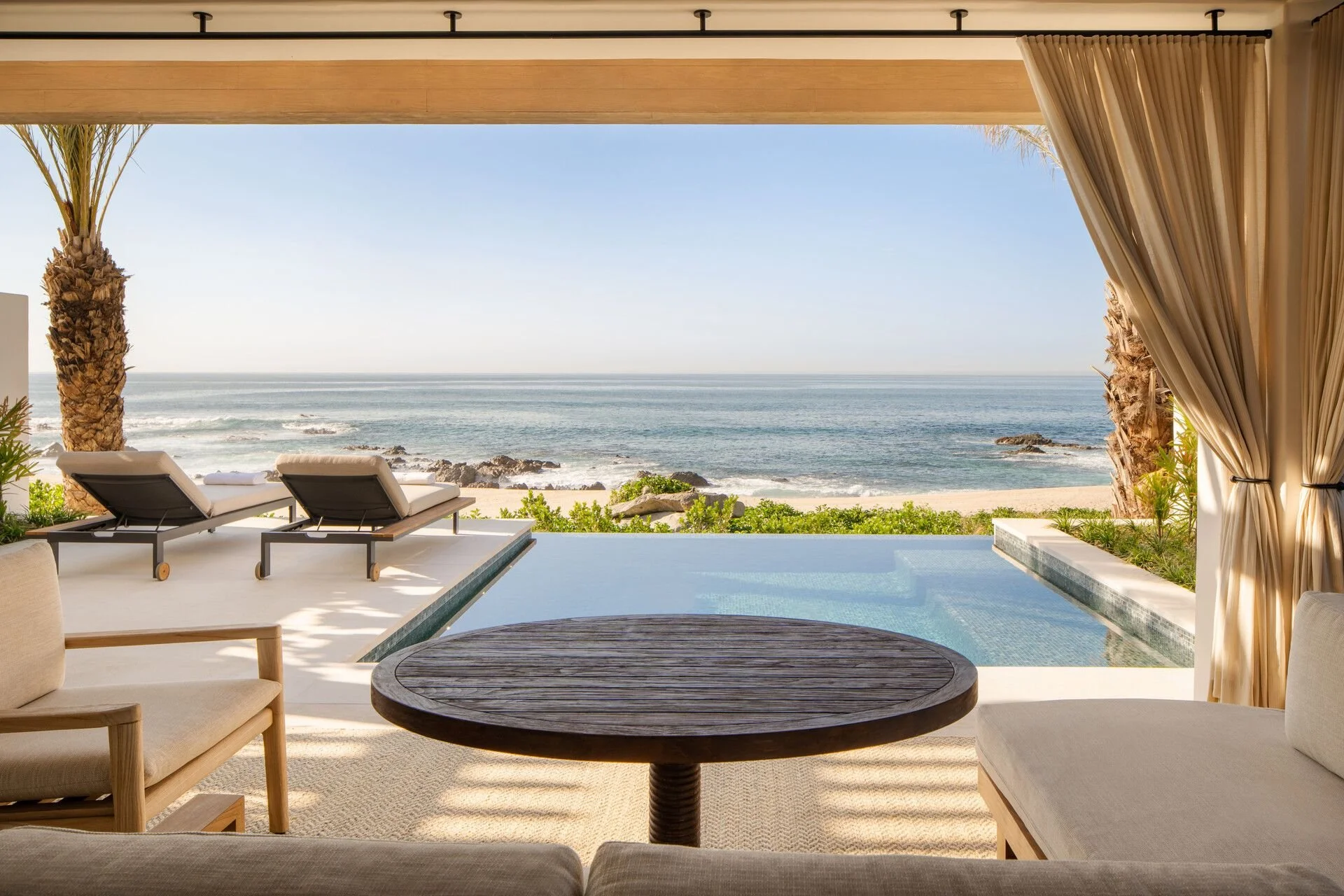 View of a modern beachfront pool area with two lounge chairs, lush greenery, and the ocean in the background, framed by beige curtains and wooden beams.