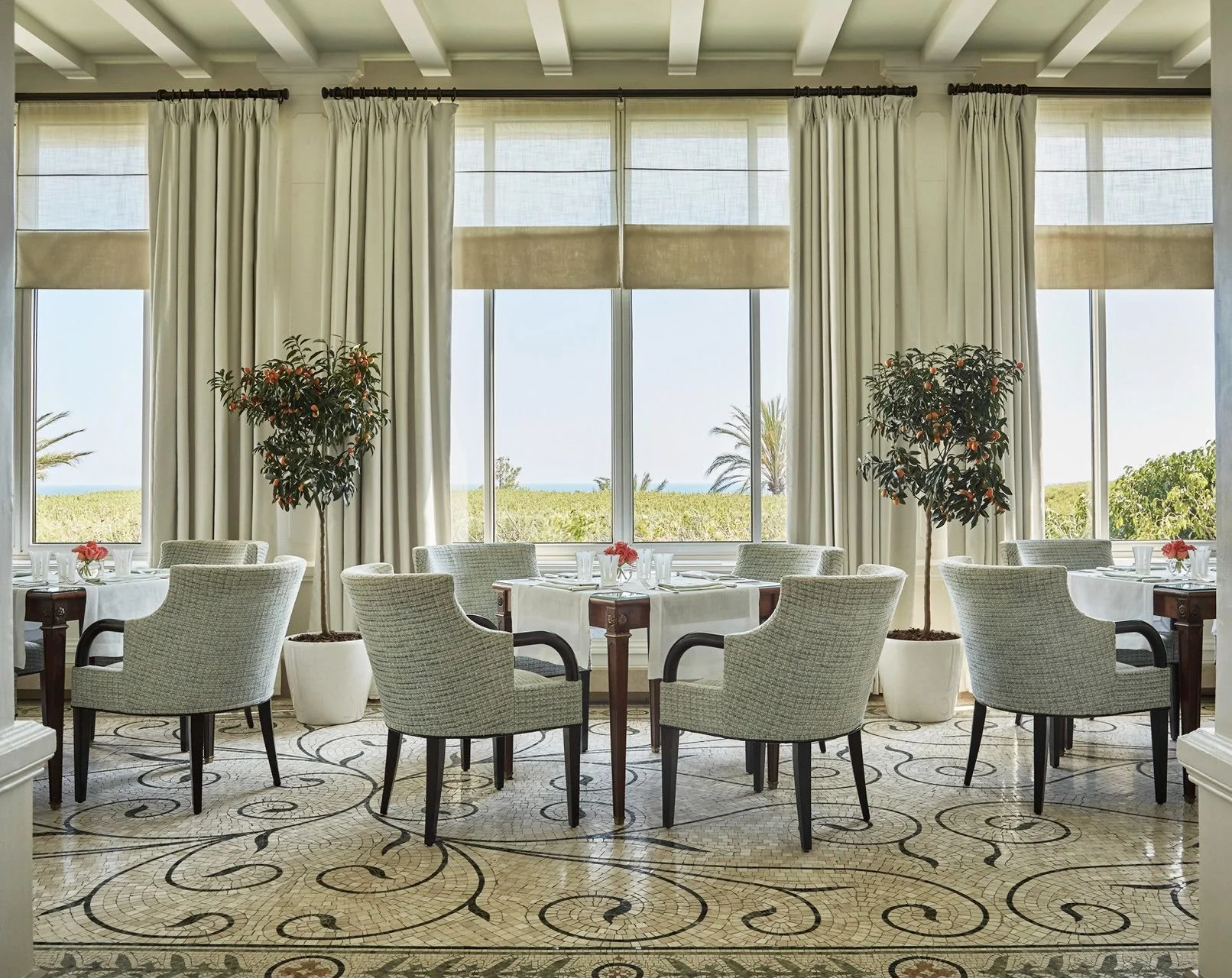 Elegant restaurant dining area with large windows, white curtains, potted orange trees, and tables set with white tablecloths and pink flowers.